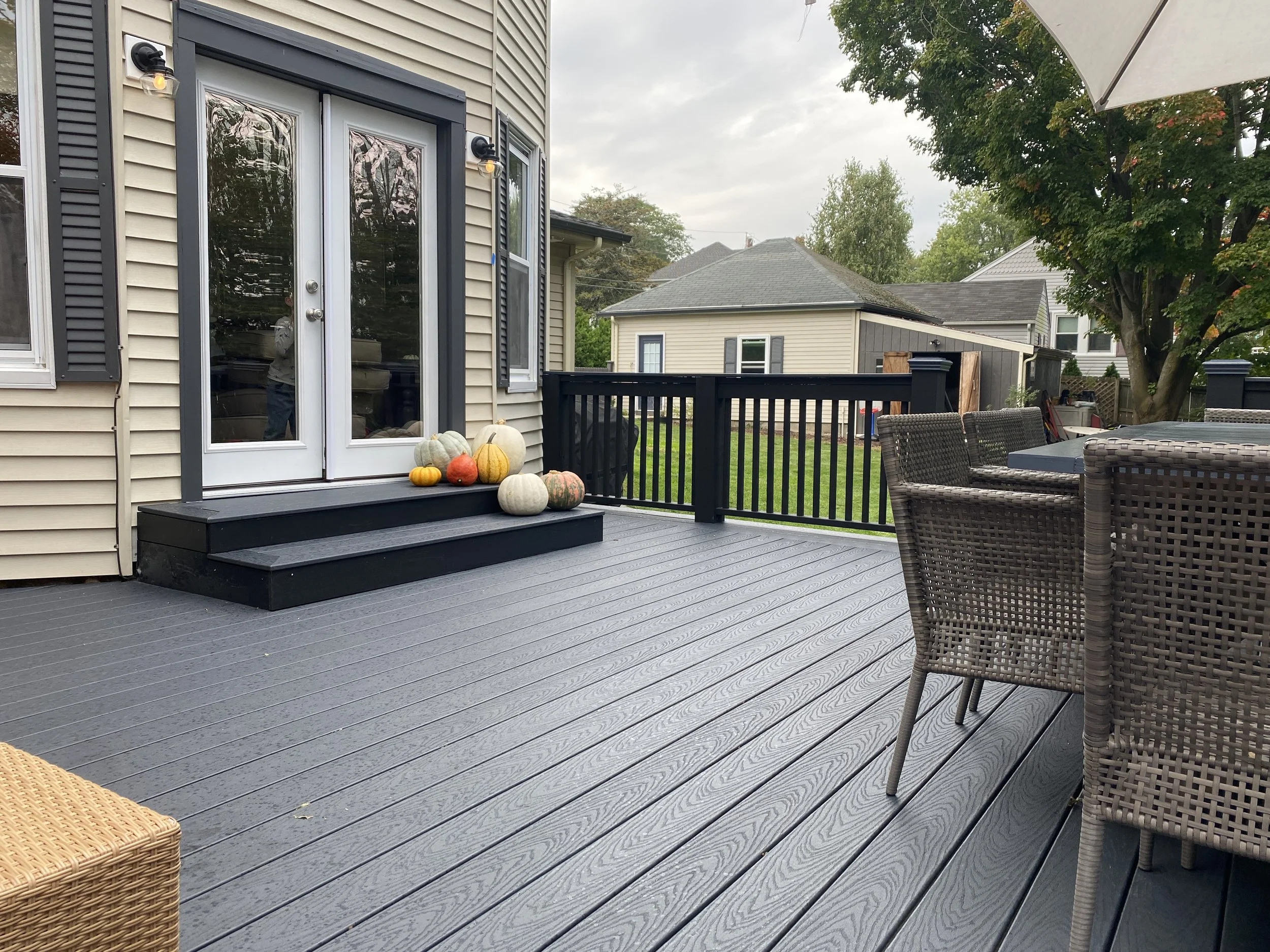 A backyard patio with a new gray deck, a black railing, and a set of black steps leading to a house with a glass door. Decorative pumpkins are arranged on the stairs. There is outdoor dining furniture, including a table with wicker chairs, and a large white umbrella providing shade. In the background, there are neighboring houses and a green yard with trees.