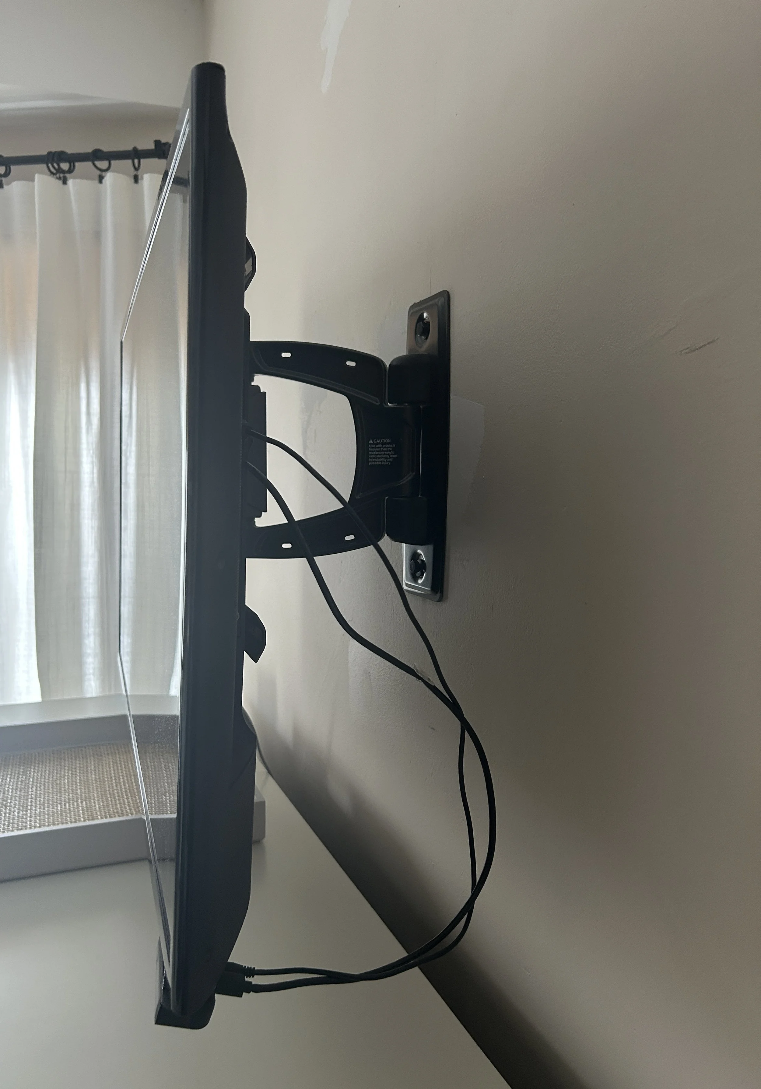 A flat-screen TV wall mount attached to a wall with two screws, with power and HDMI cables hanging down, near a window with light-colored curtains.