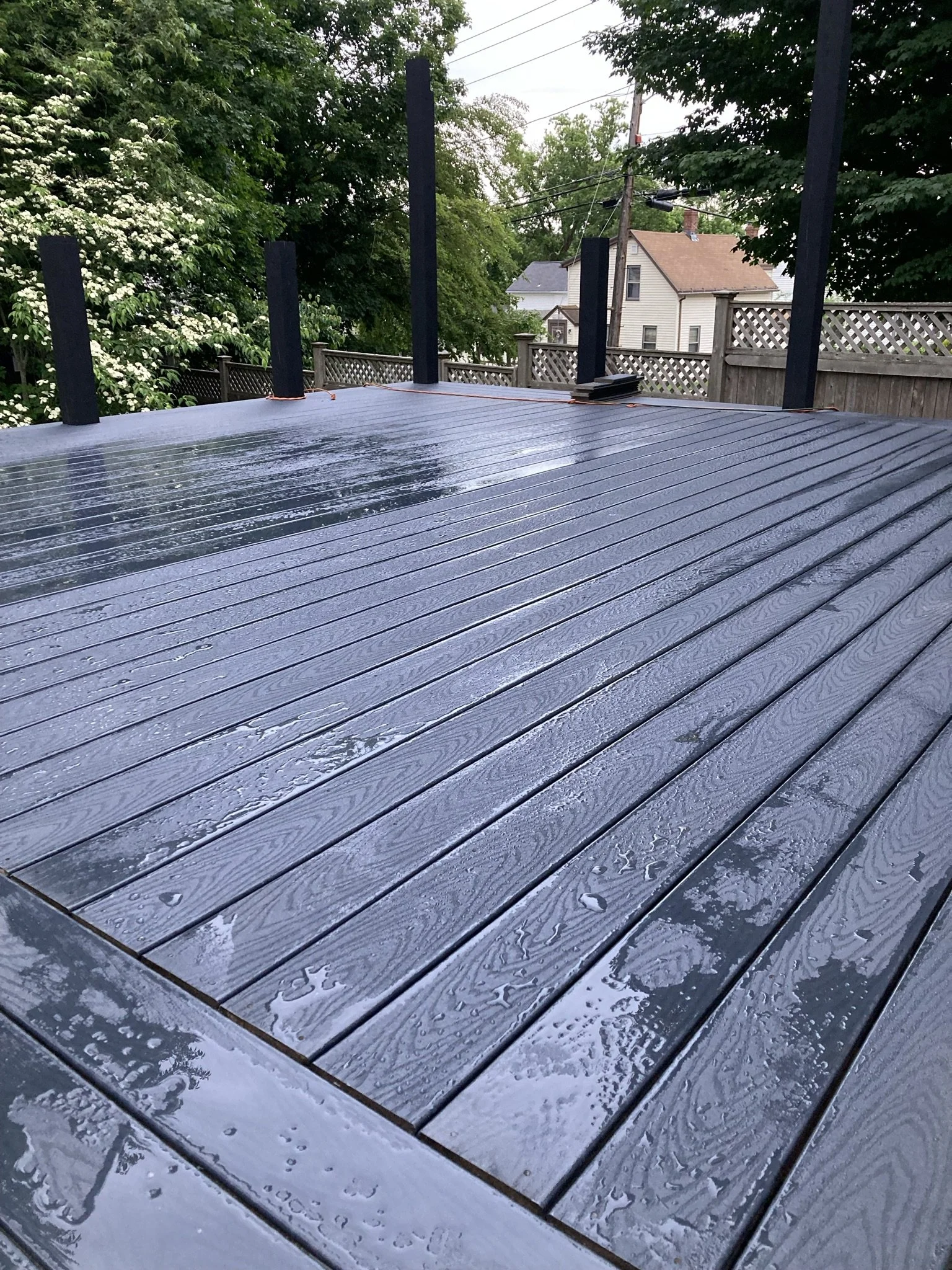 Wet, newly painted dark gray wooden deck in a backyard with green trees and neighboring houses.