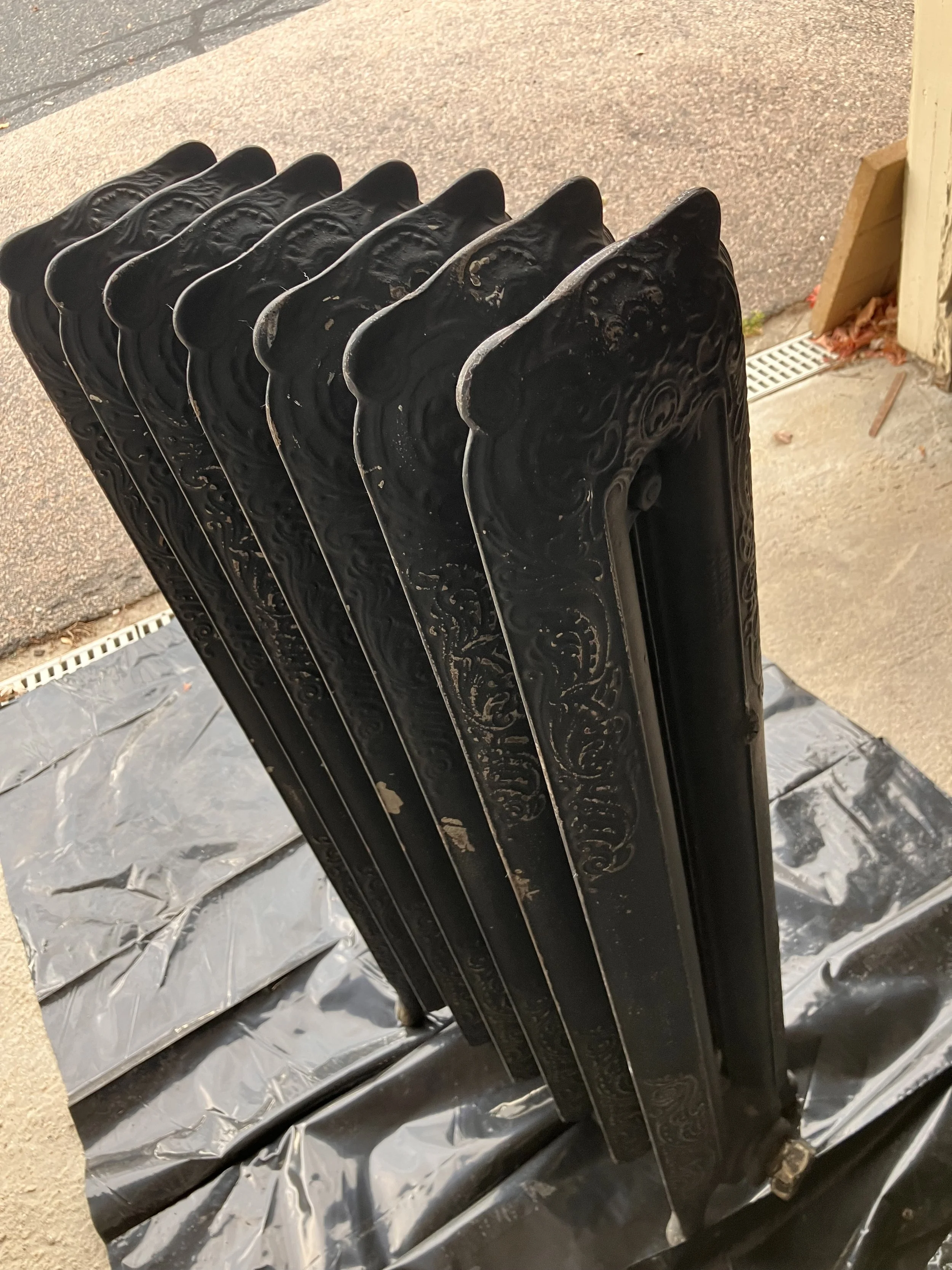A black cast iron outdoor radiator with ornate, decorative patterns, placed on a black plastic sheet on a concrete sidewalk.