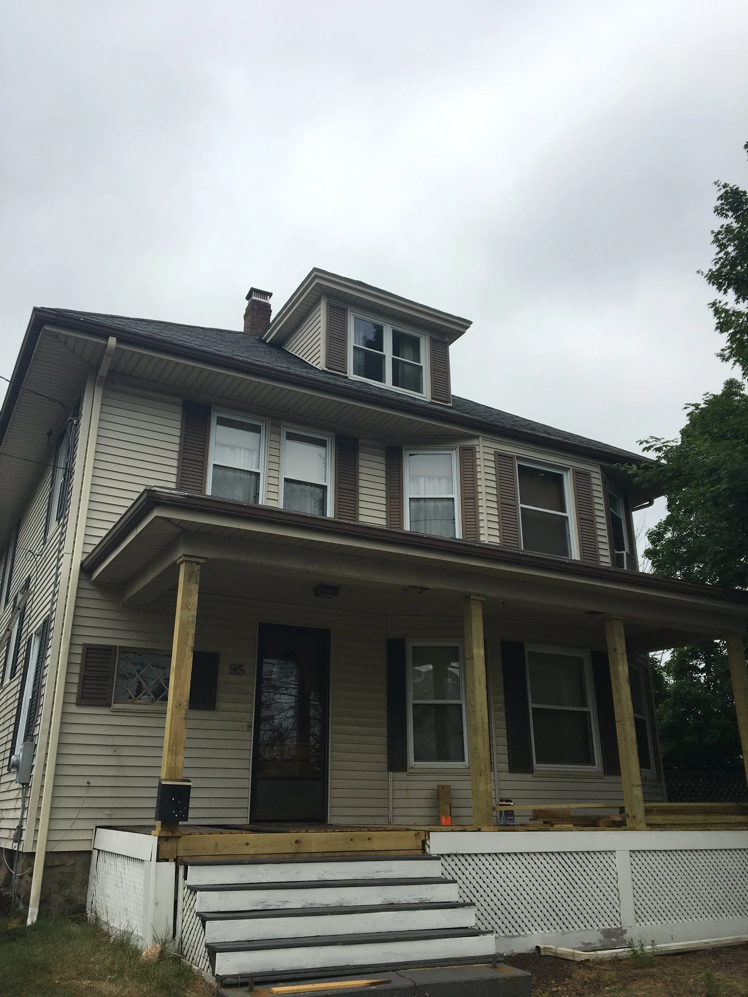 A multi-story house under construction, with a porch supported by wooden posts, stairs leading up to the entrance, and new deck flooring being installed.