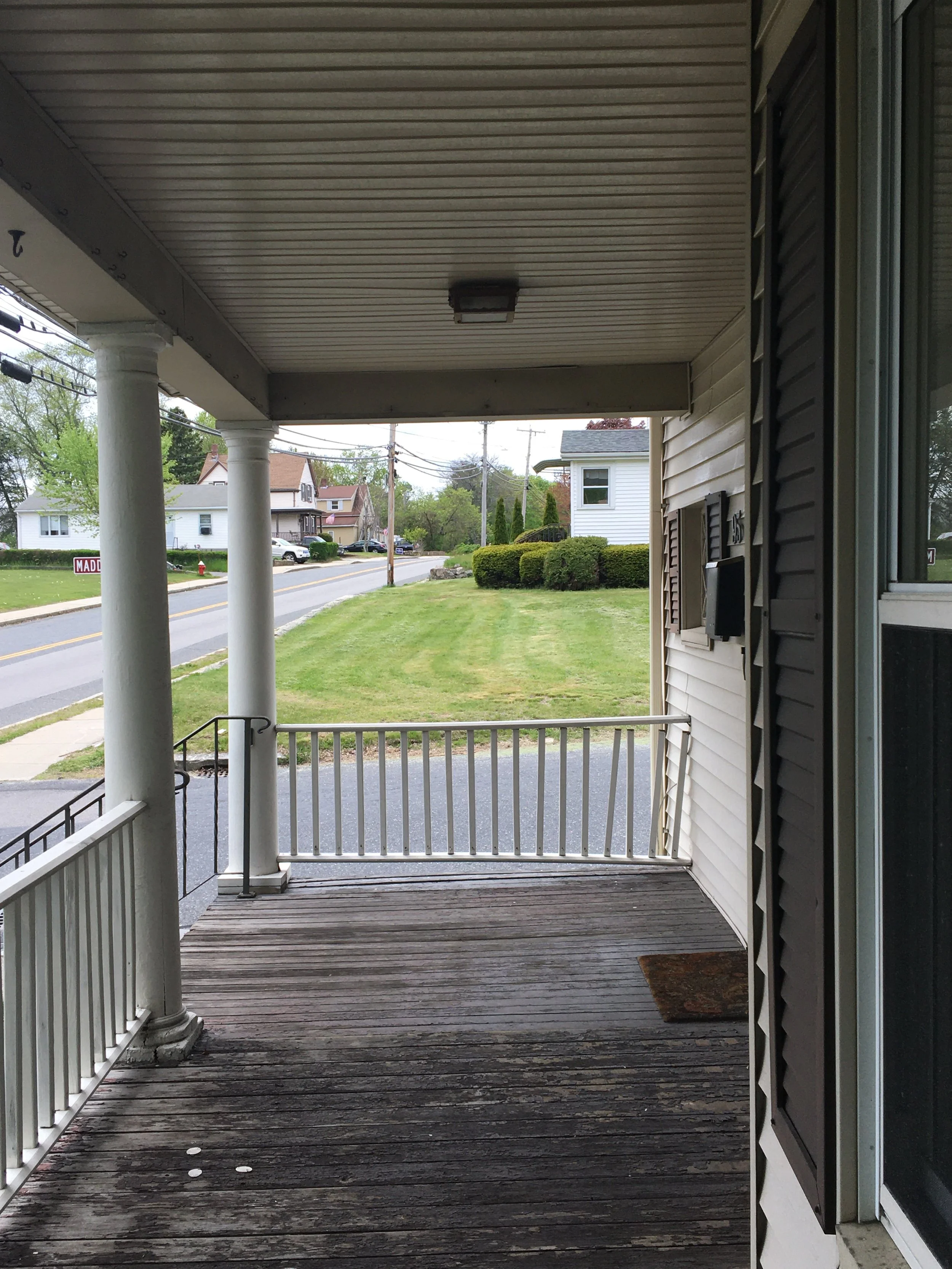View of a front porch with wooden flooring, white railing, and columns, overlooking a grassy yard with bushes and a street with houses in the background.