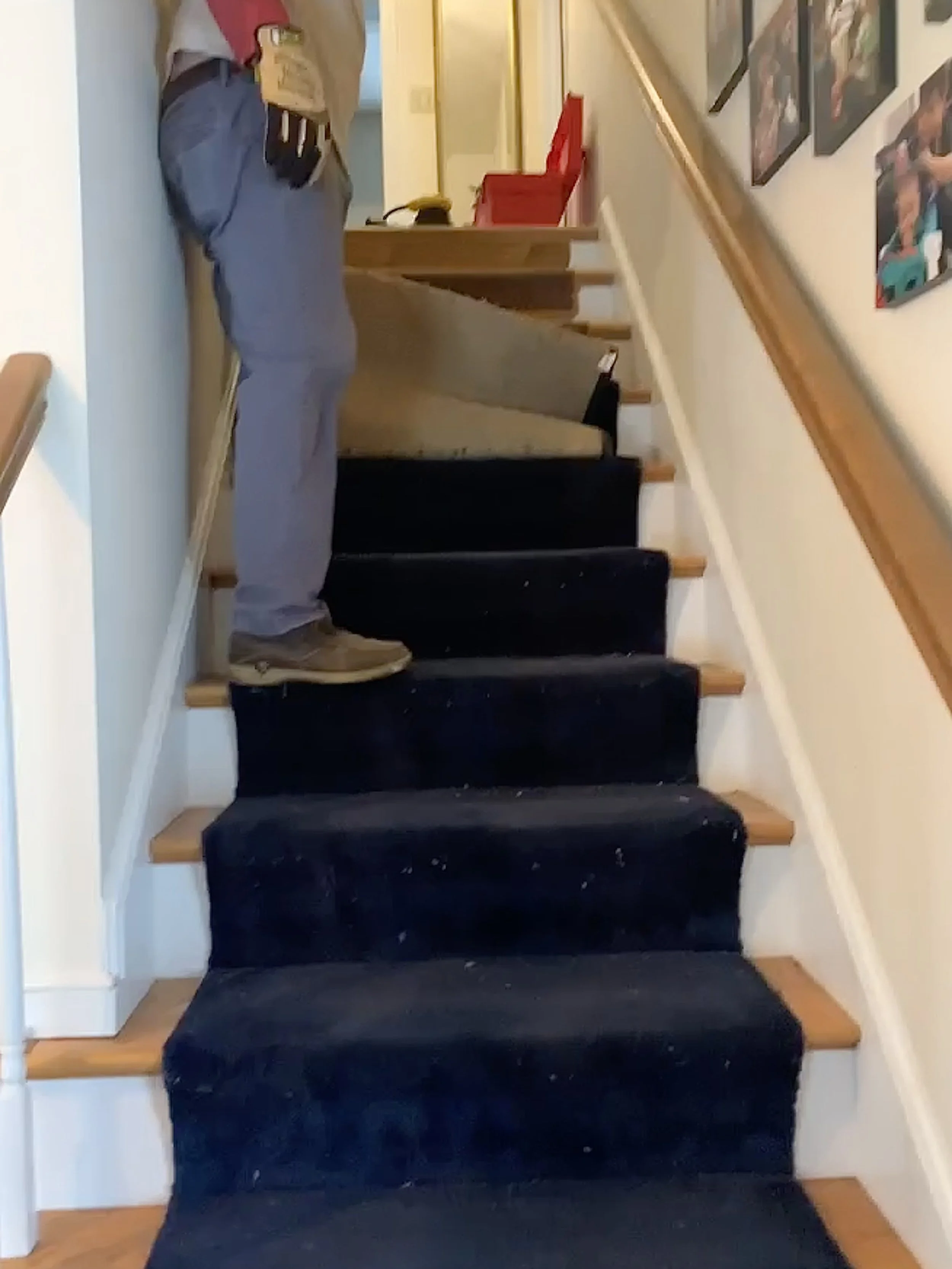 A person standing on a staircase with dark carpeted steps and wooden edges, near a wall decorated with framed photos.