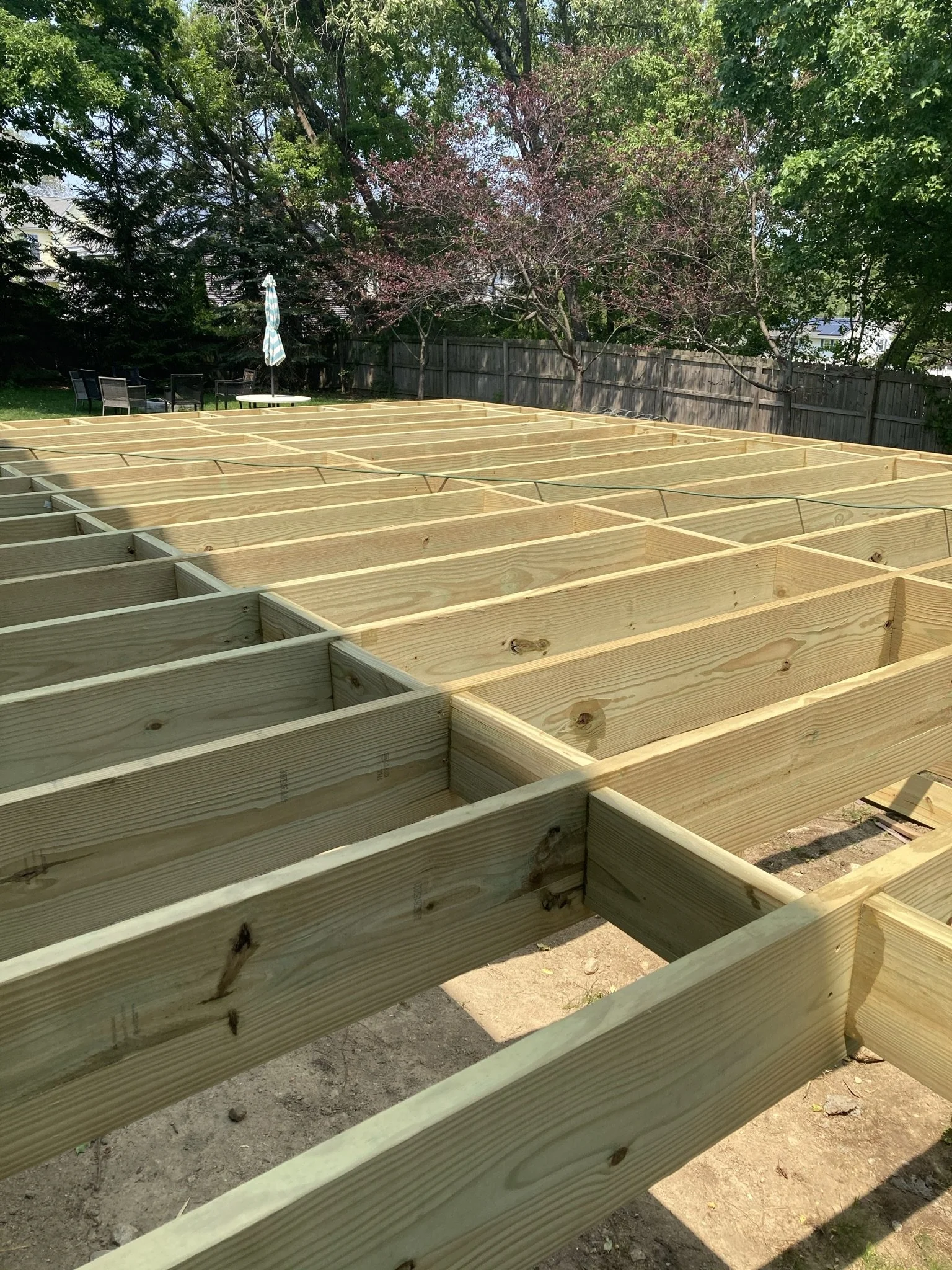 Wooden framing for a deck being constructed in a backyard, with trees and outdoor furniture in the background.