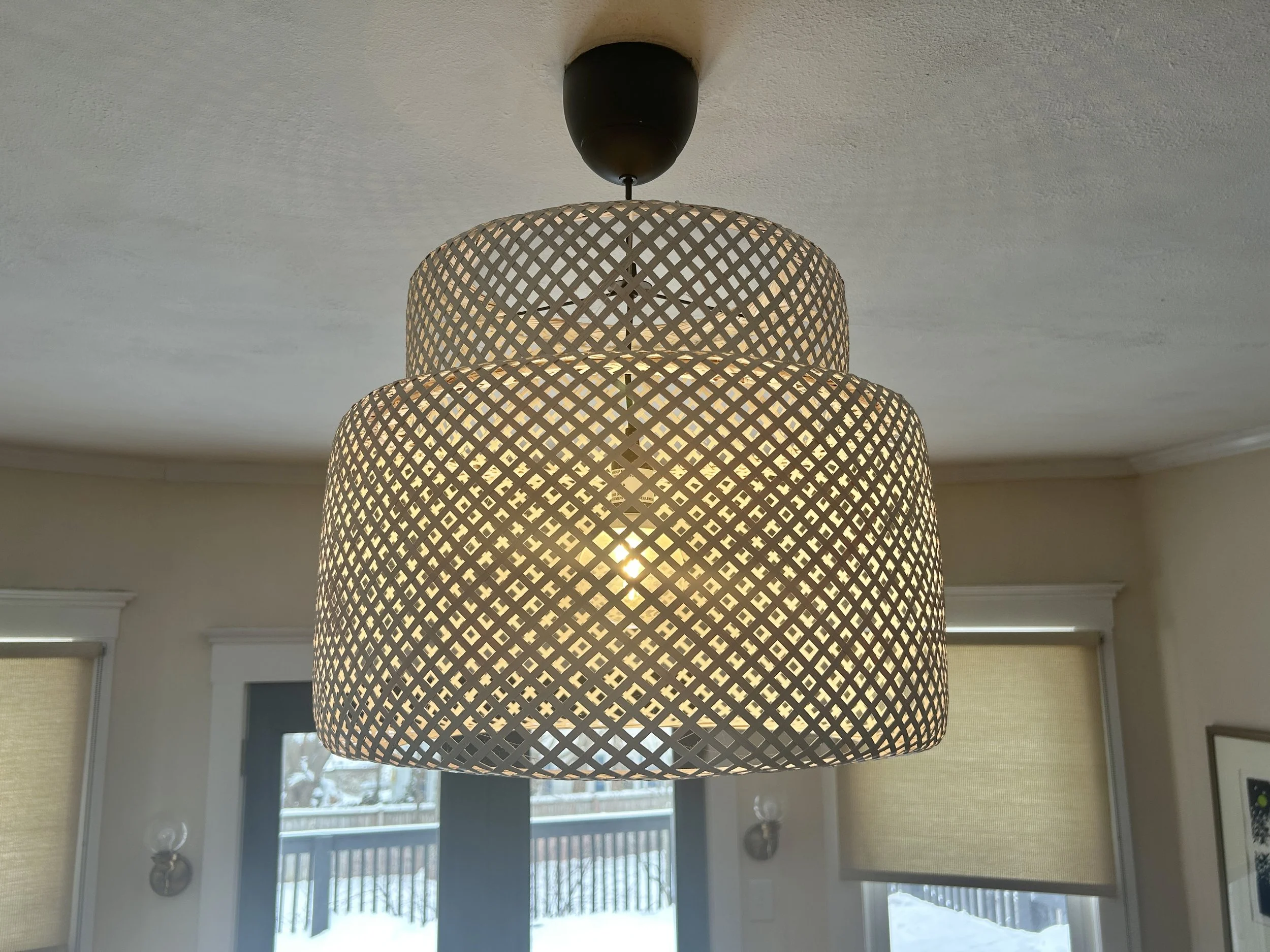 Large woven pendant light fixture hanging from the ceiling in a room with windows showing a snowy outdoor scene.