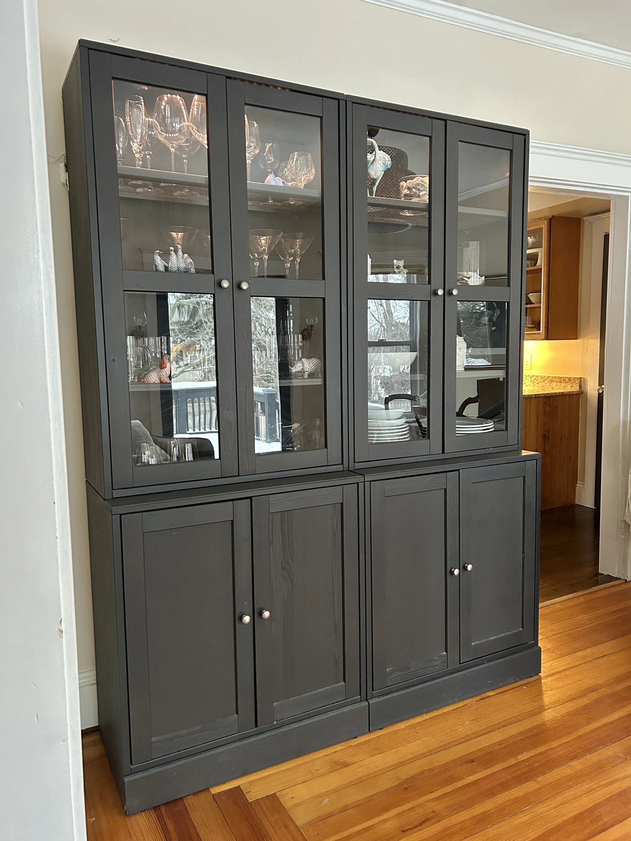 A black wooden china cabinet with glass doors displaying glassware and decorative figurines, placed against a white wall with hardwood floors in a home interior.