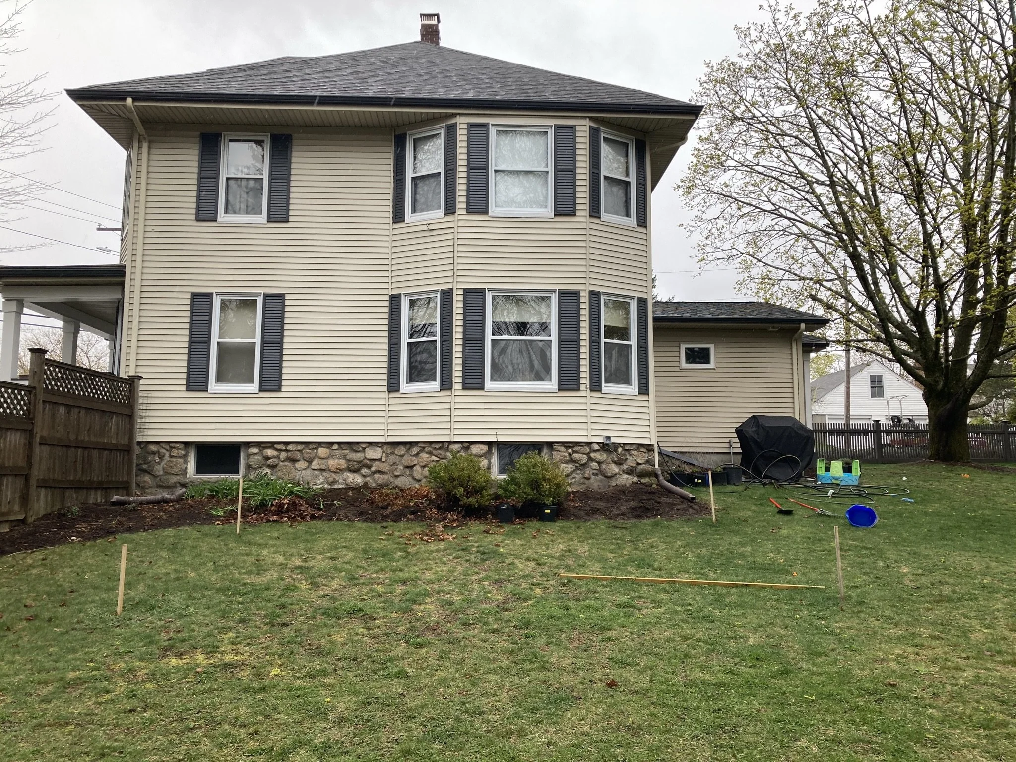 A two-story house with beige siding and blue shutters has a garden in front and a large tree on the right. There are gardening tools and equipment, including a black grill, a blue sled, and hoses, scattered on the lawn, indicating yard work in progre