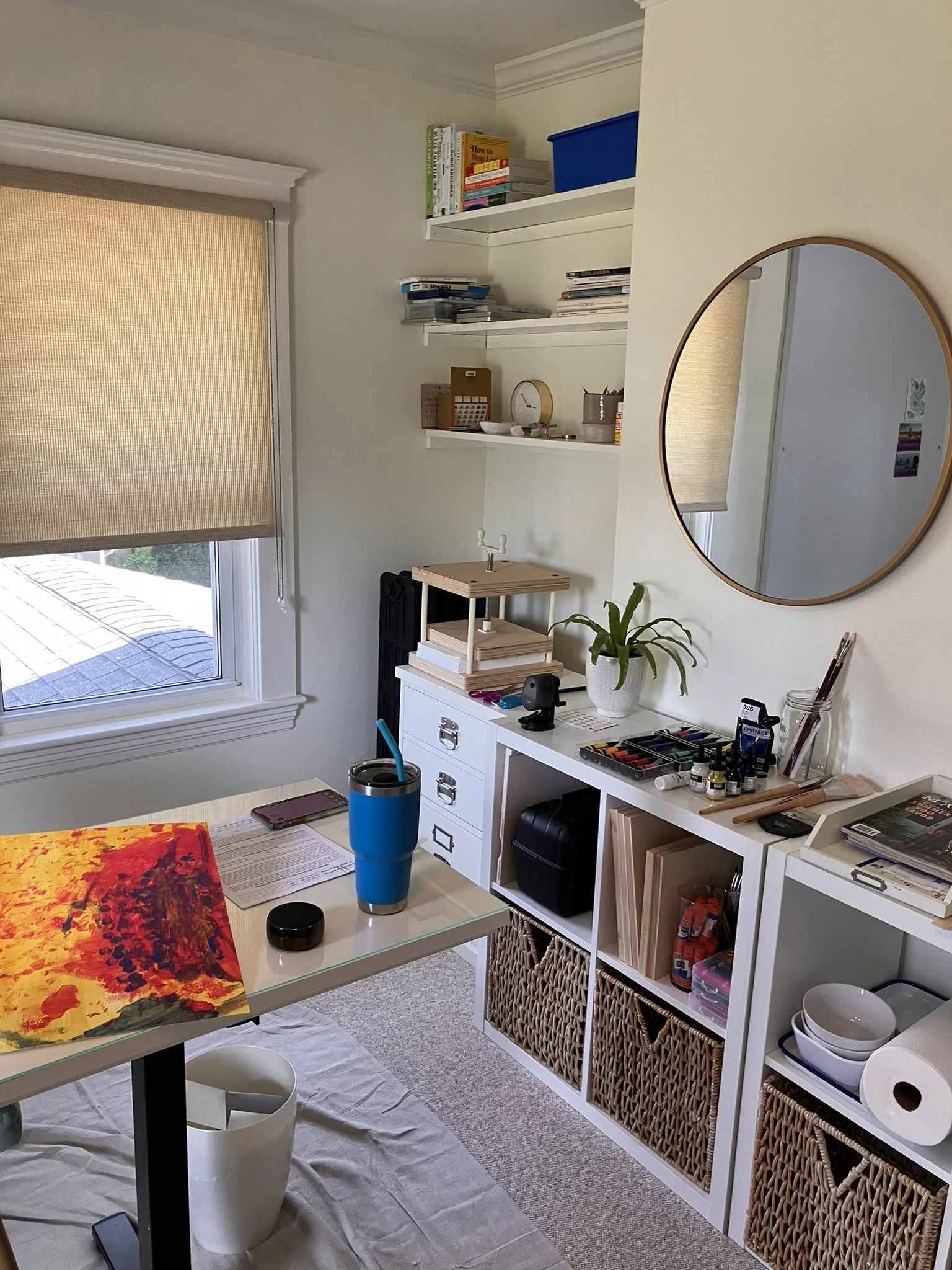 A room with a window on the left, with beige blinds. A white desk with colorful artwork, a blue tumbler, and various papers is in the foreground. White shelving units hold books, storage bins, and art supplies. A mirror hangs on the right wall, refle