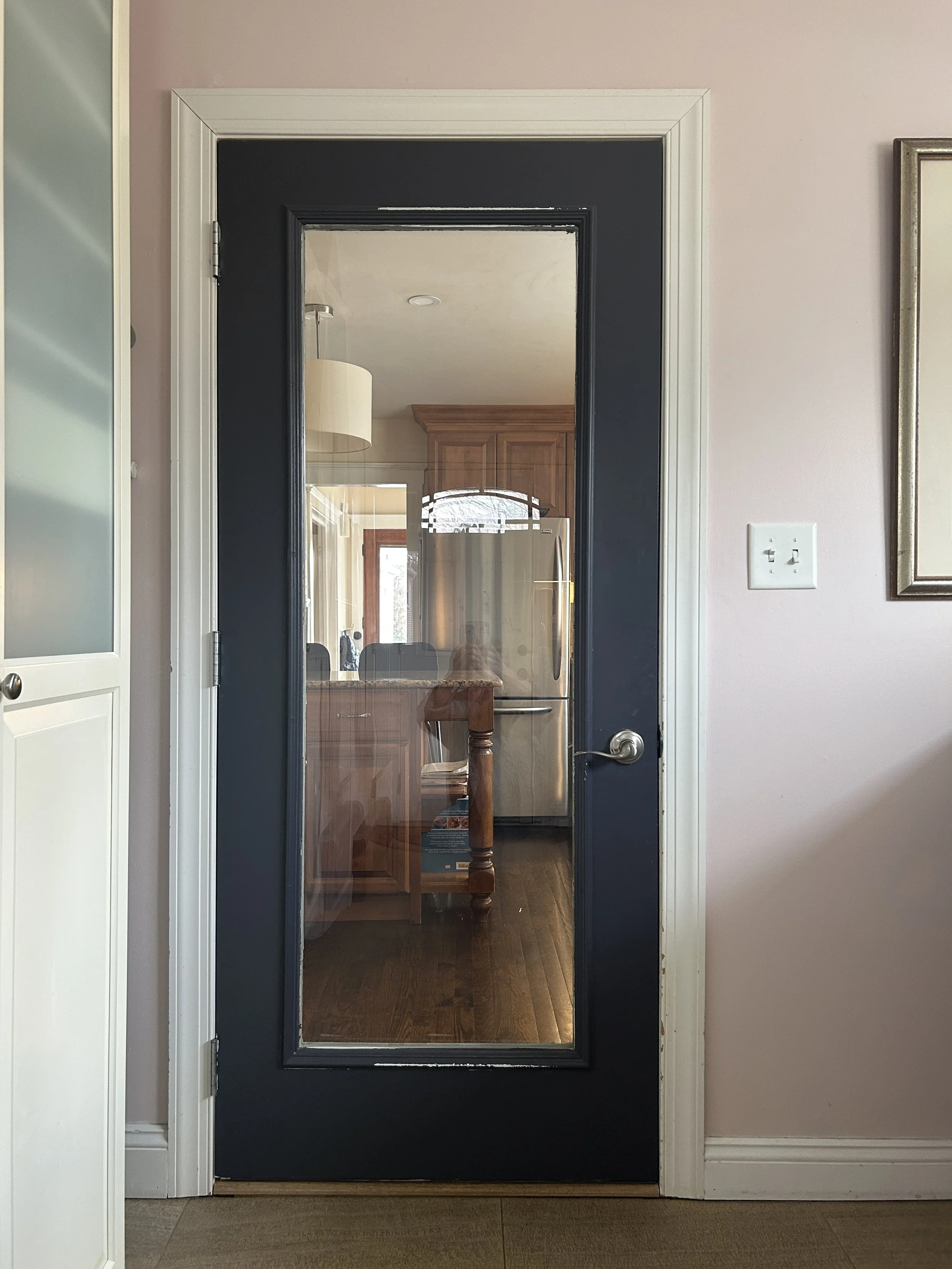 View of a black interior door with a large glass panel, leading to a kitchen area with wooden cabinets and stainless steel refrigerator.