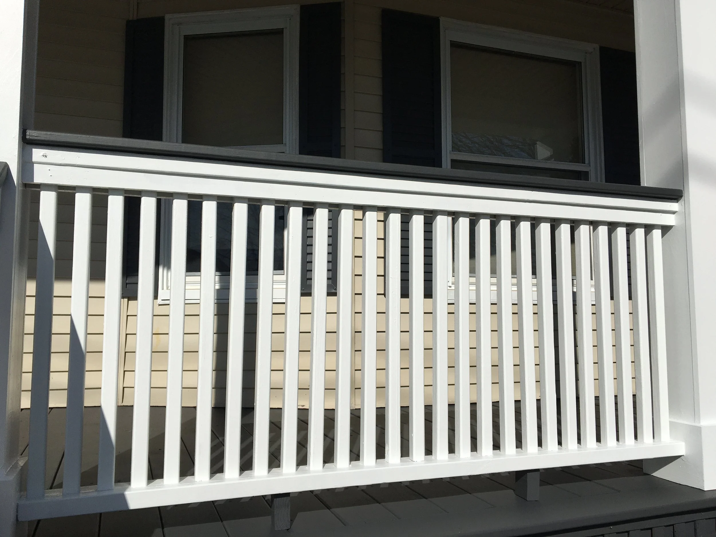 A white balcony railing in front of beige-colored house siding and two window sections, with some shadows cast on it.