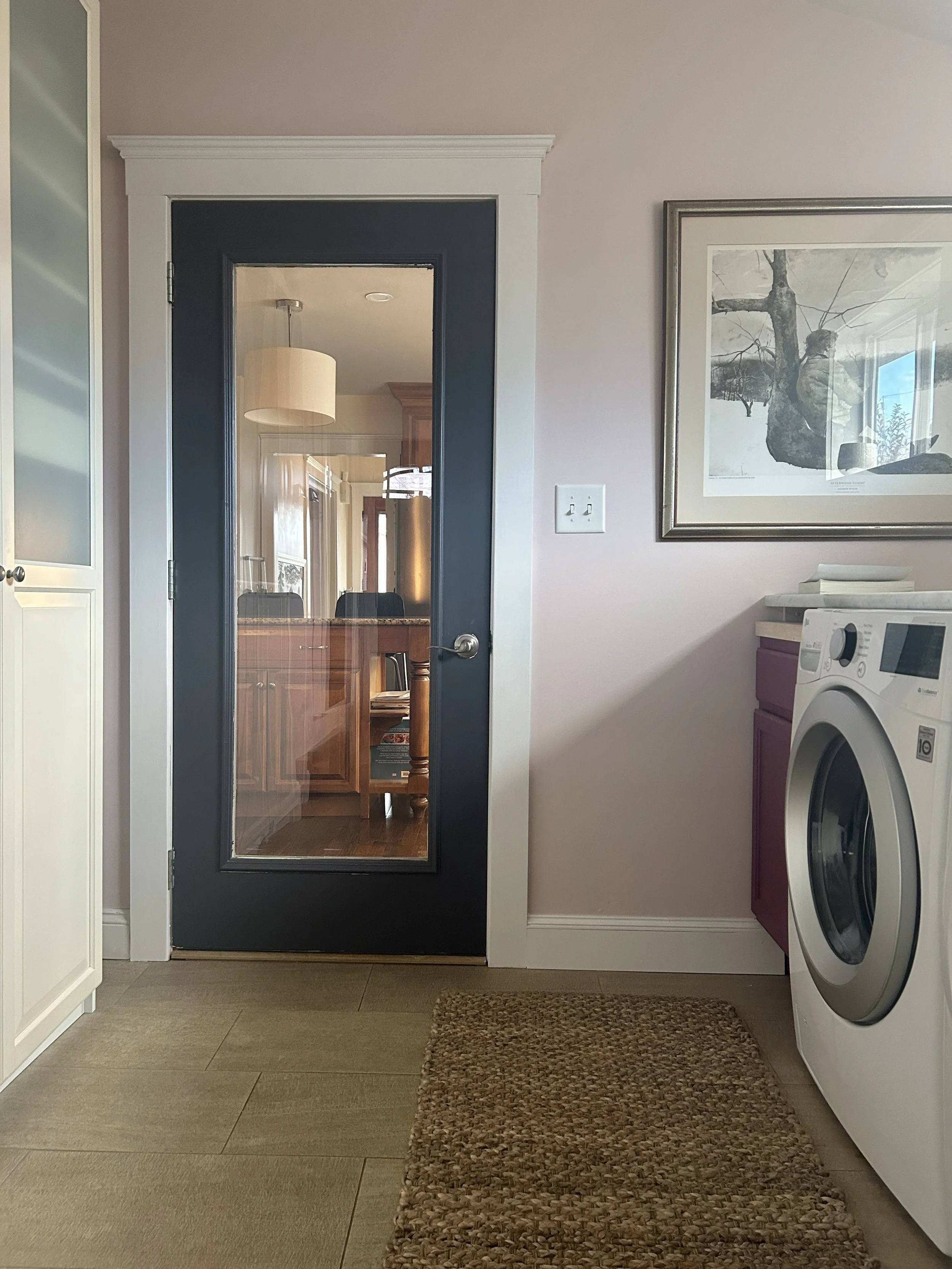 Interior view of a laundry room with a front-loading washing machine on the right, a woven brown rug on beige tile flooring, a white framed glass door leading to a dining area, beige walls with a framed black-and-white tree picture, and part of a whi