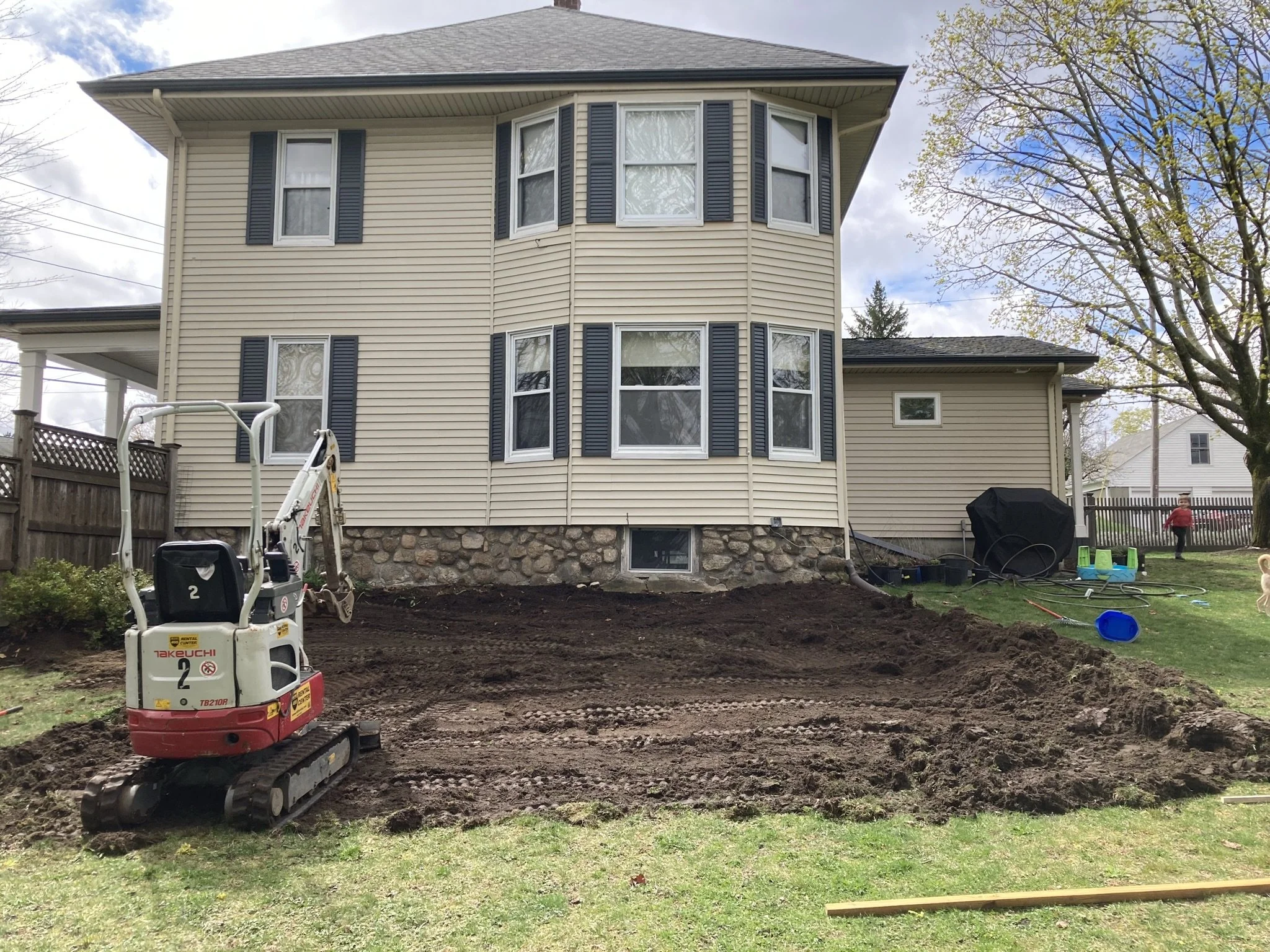 A backyard with a house under renovation, featuring a small excavator on freshly dug soil area, and children's play equipment on the grass, with a tree and neighboring house in the background.