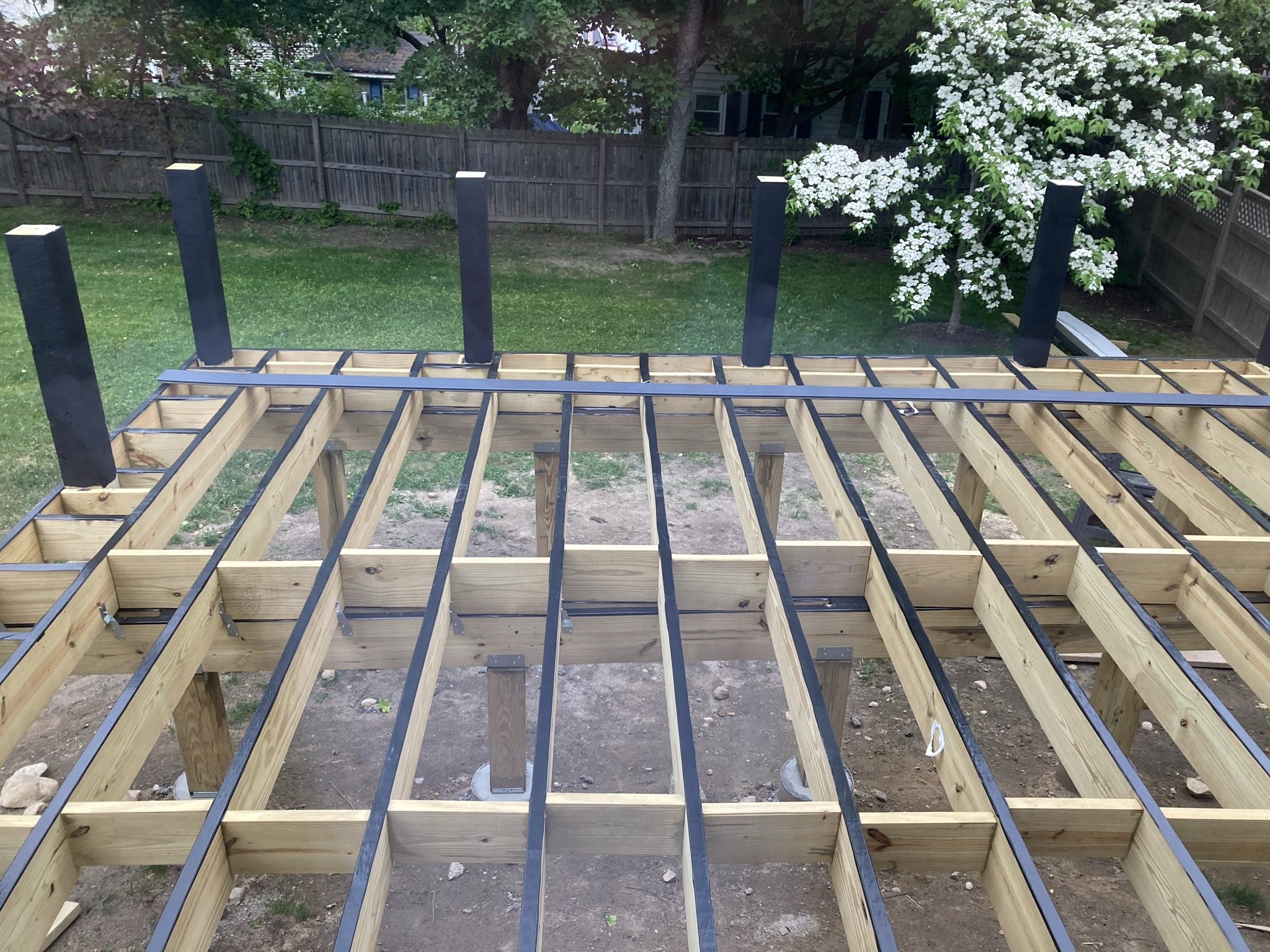 Under-construction wooden deck framework with black metal beams in a backyard with grass, trees, and a flowering bush.