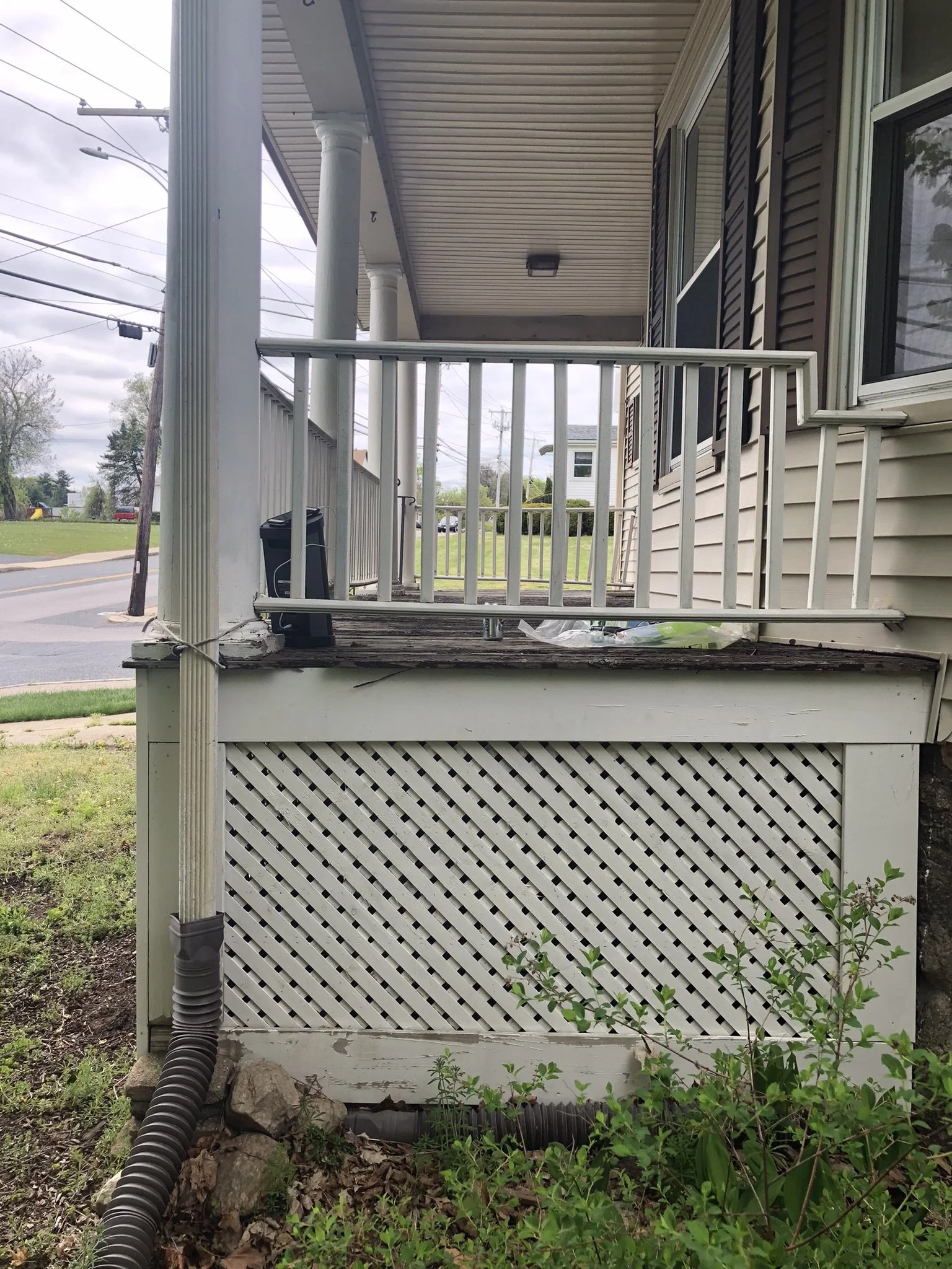 View of a house's front porch with white railings and columns, a black doormat, and a lattice foundation, with some overgrown plants and a black drainage pipe on the ground.