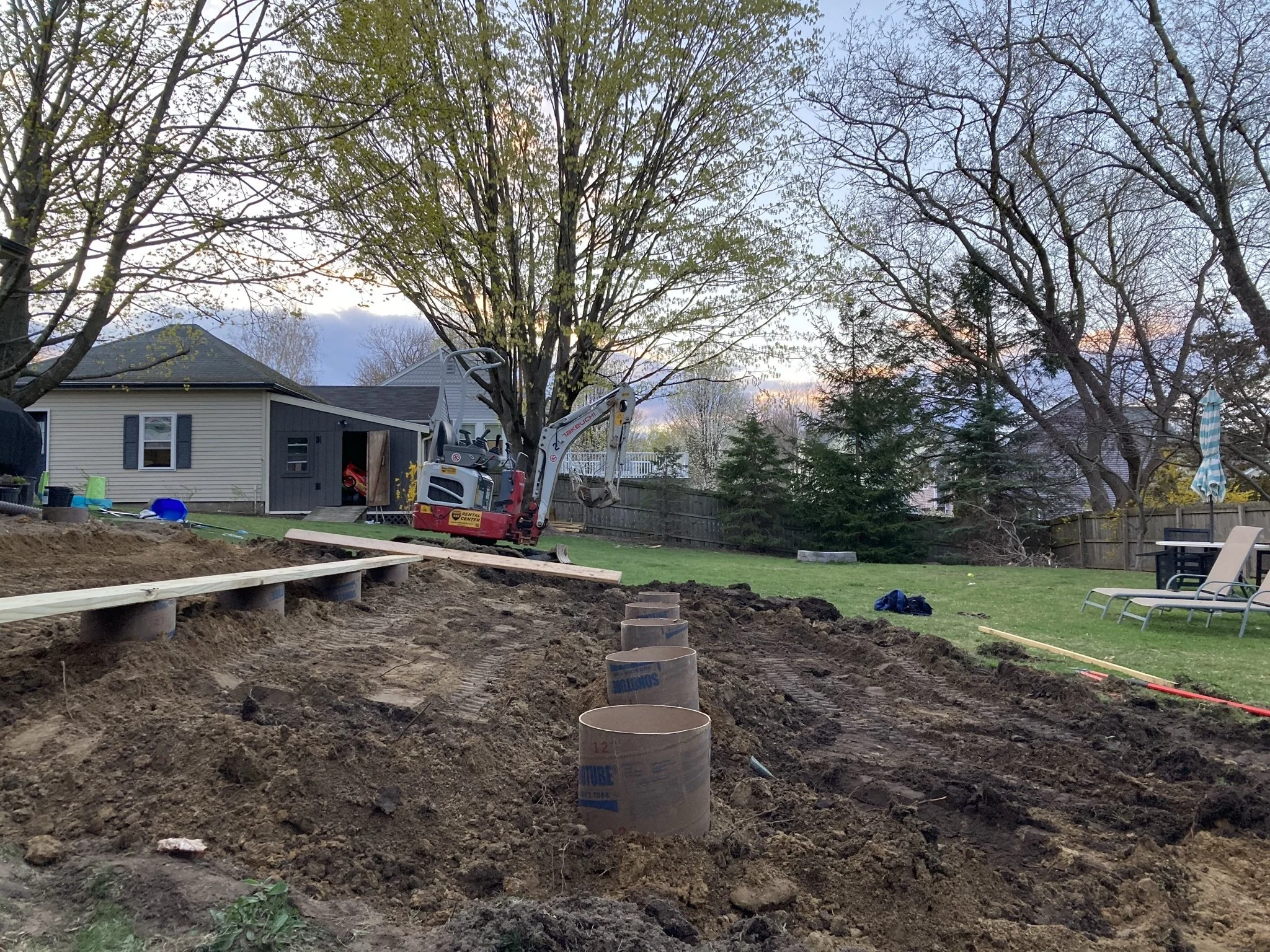 A backyard under construction with a mini excavator, exposed pipes, and a wooden framework, with trees, grass, and outdoor furniture in the background.