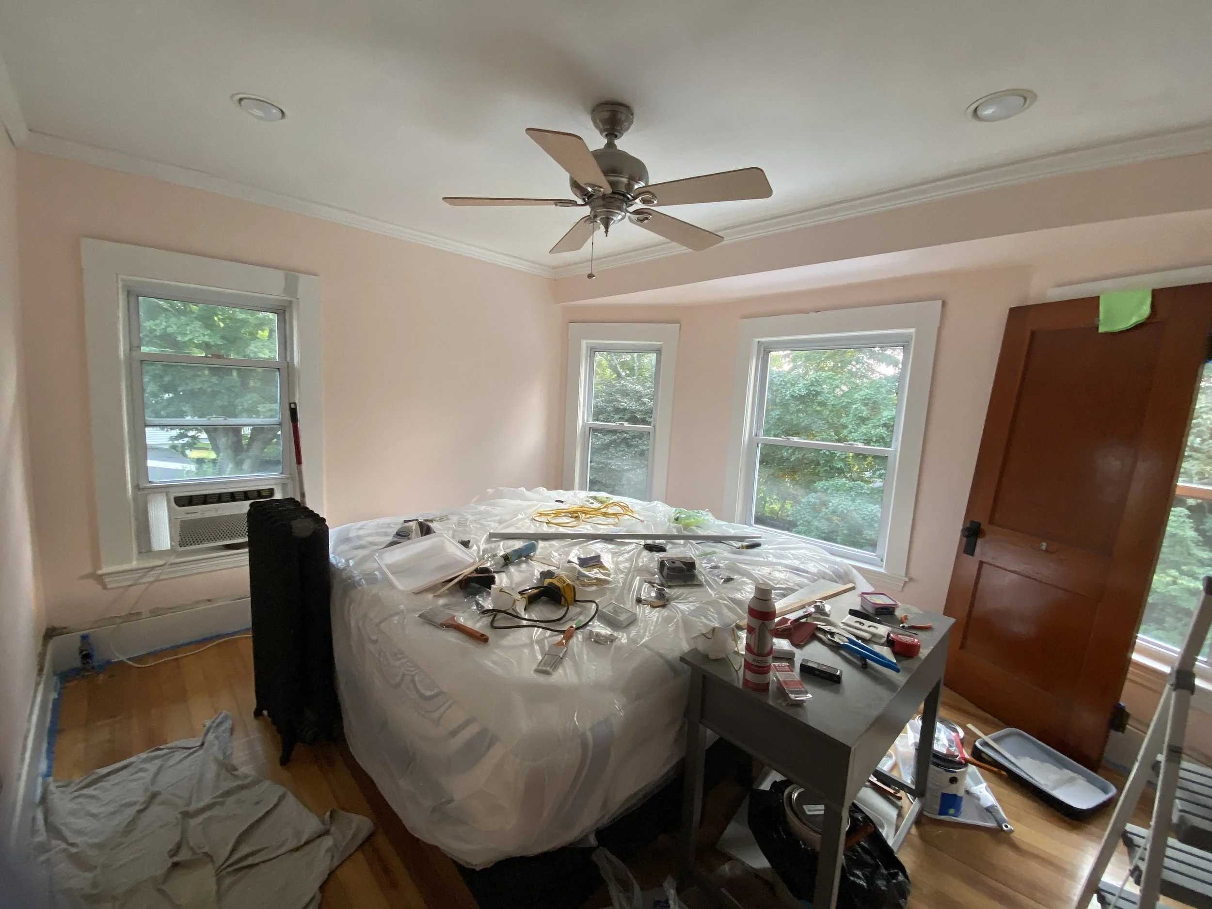 Room with pink walls, a ceiling fan, three windows, a bed covered with plastic and various tools and supplies on top, a small table nearby with more supplies, and an open wooden door.