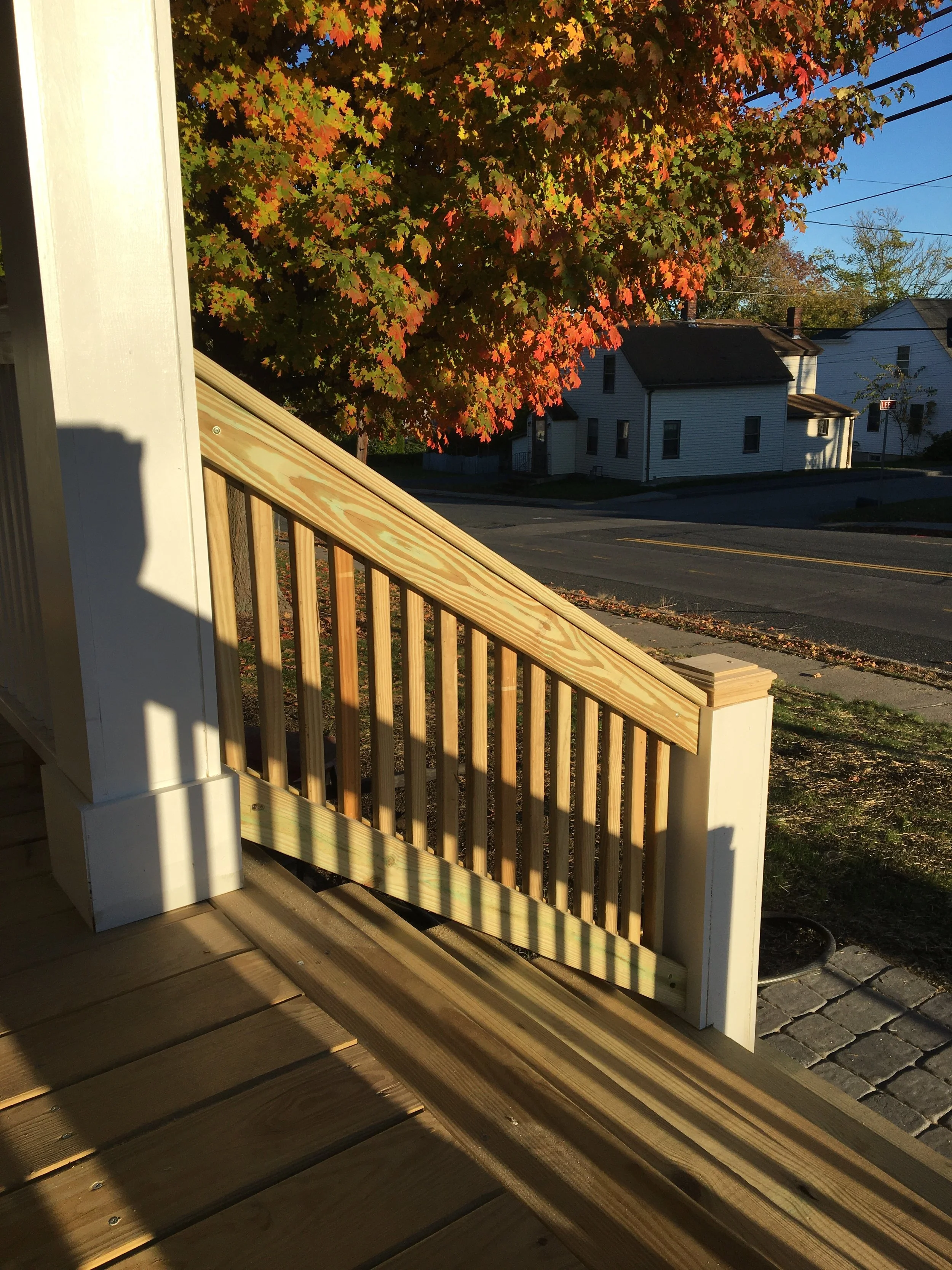 View of a newly built wooden porch railing and stairs on a house during sunset, with a large autumn tree with red and green leaves in the background.