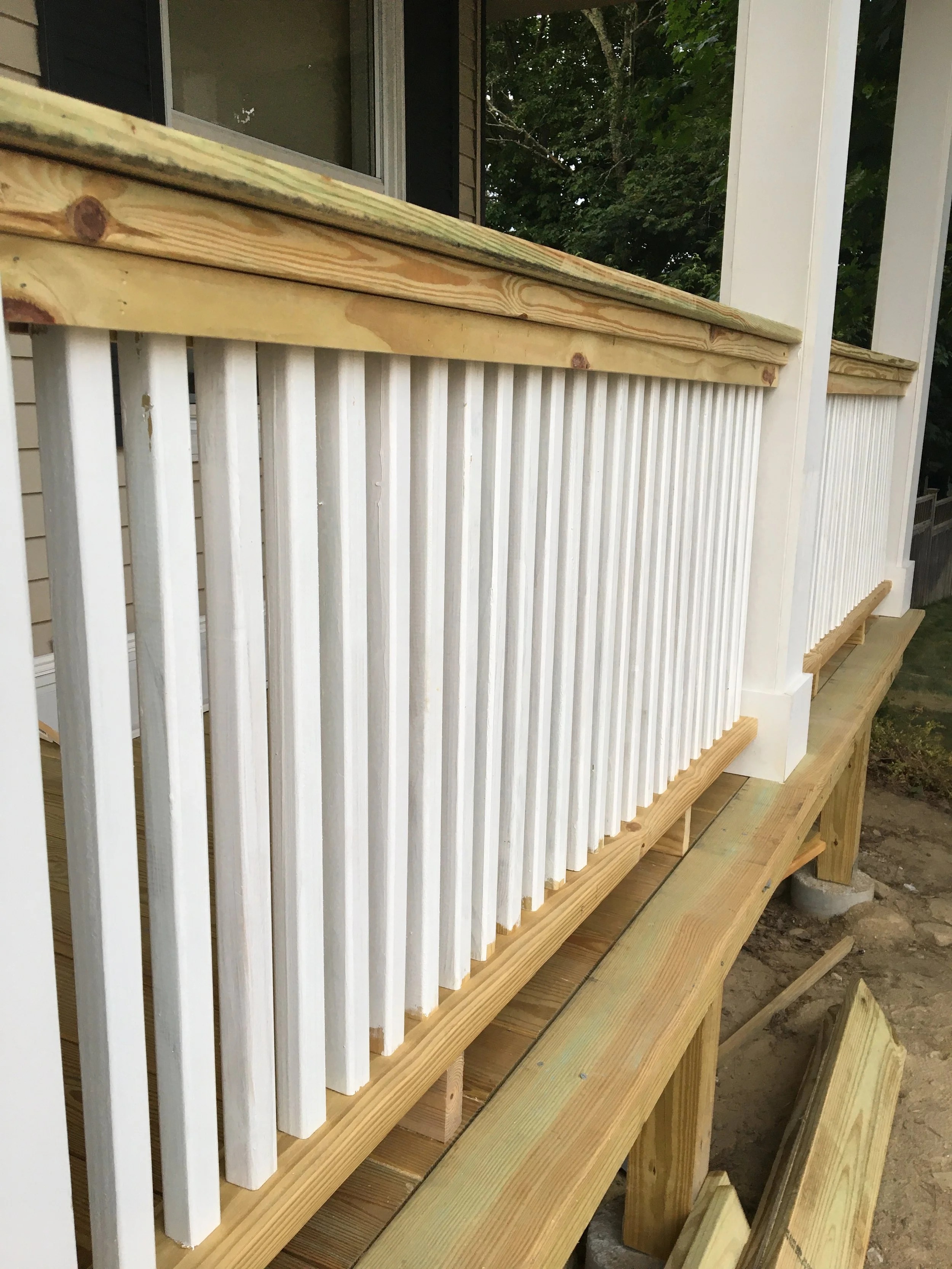 Close-up of a new wooden porch railing under construction, featuring white vertical balusters and natural wood top and bottom rails, with a house and trees in the background.