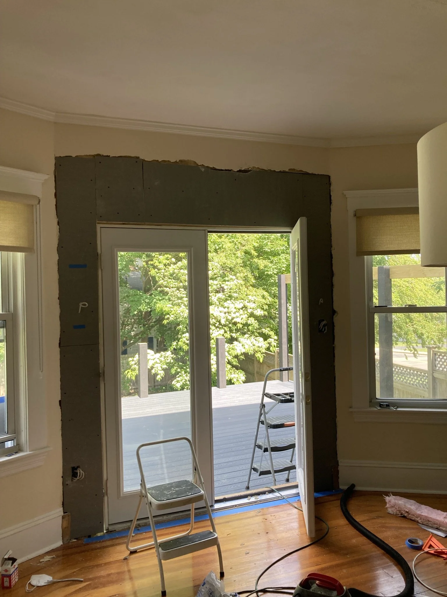 Interior of a room under renovation with an open glass door leading to a deck and trees outside, construction tools and materials scattered on the floor.