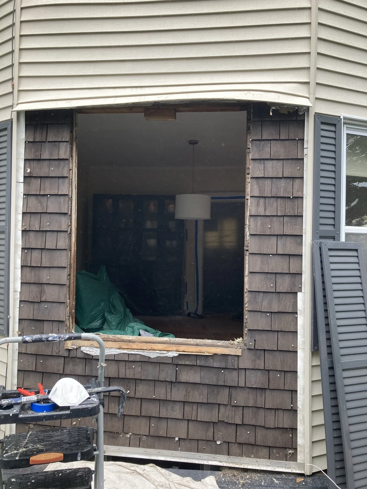 A house undergoing renovation with a large window opening, showing a room inside with a hanging pendant light and construction materials, including green bags and tools, outside scaffolding and window shutters.