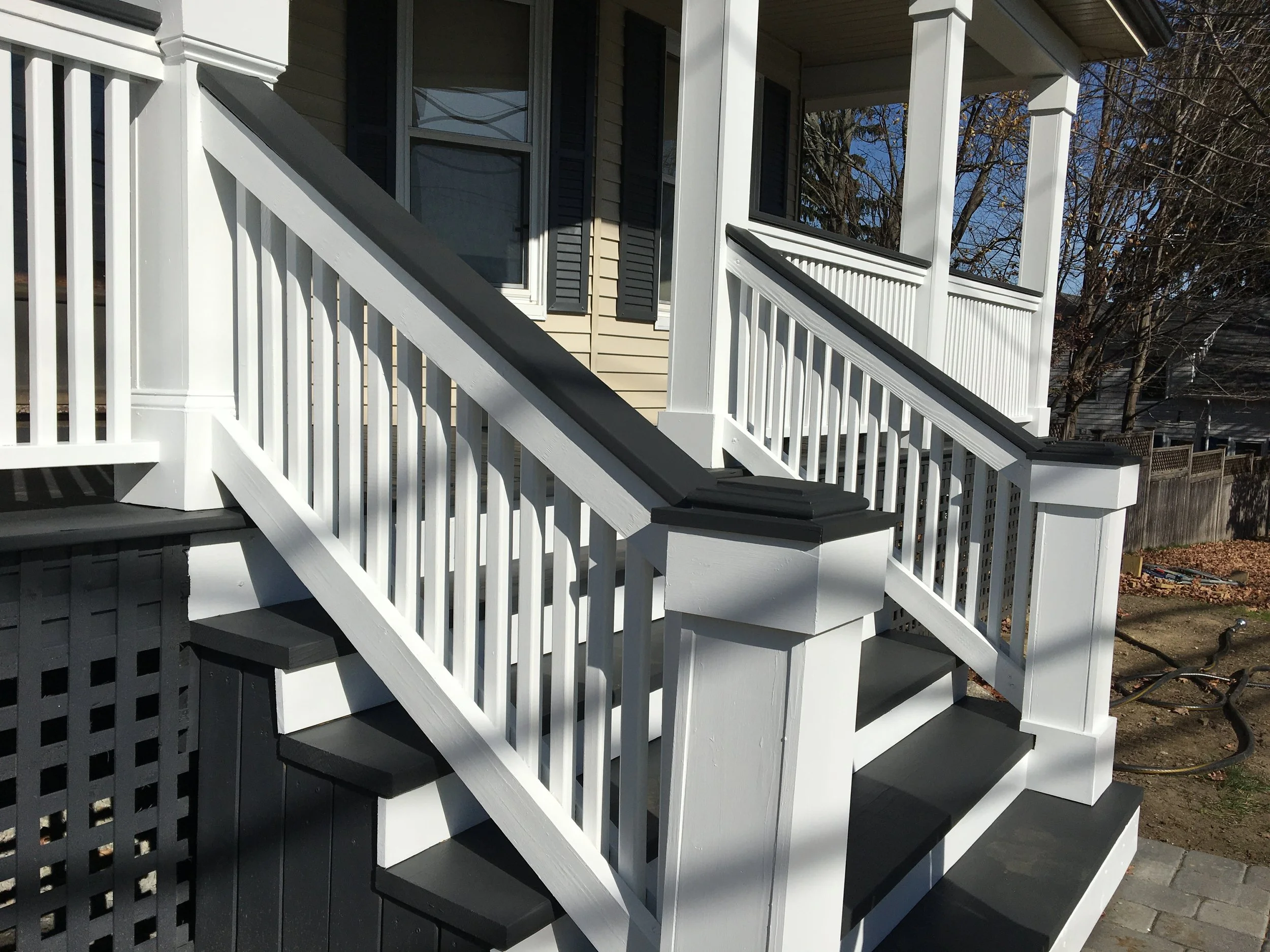 A close-up view of a white wooden porch staircase with black treads and white railings on either side, attached to a house with beige siding and black window shutters.