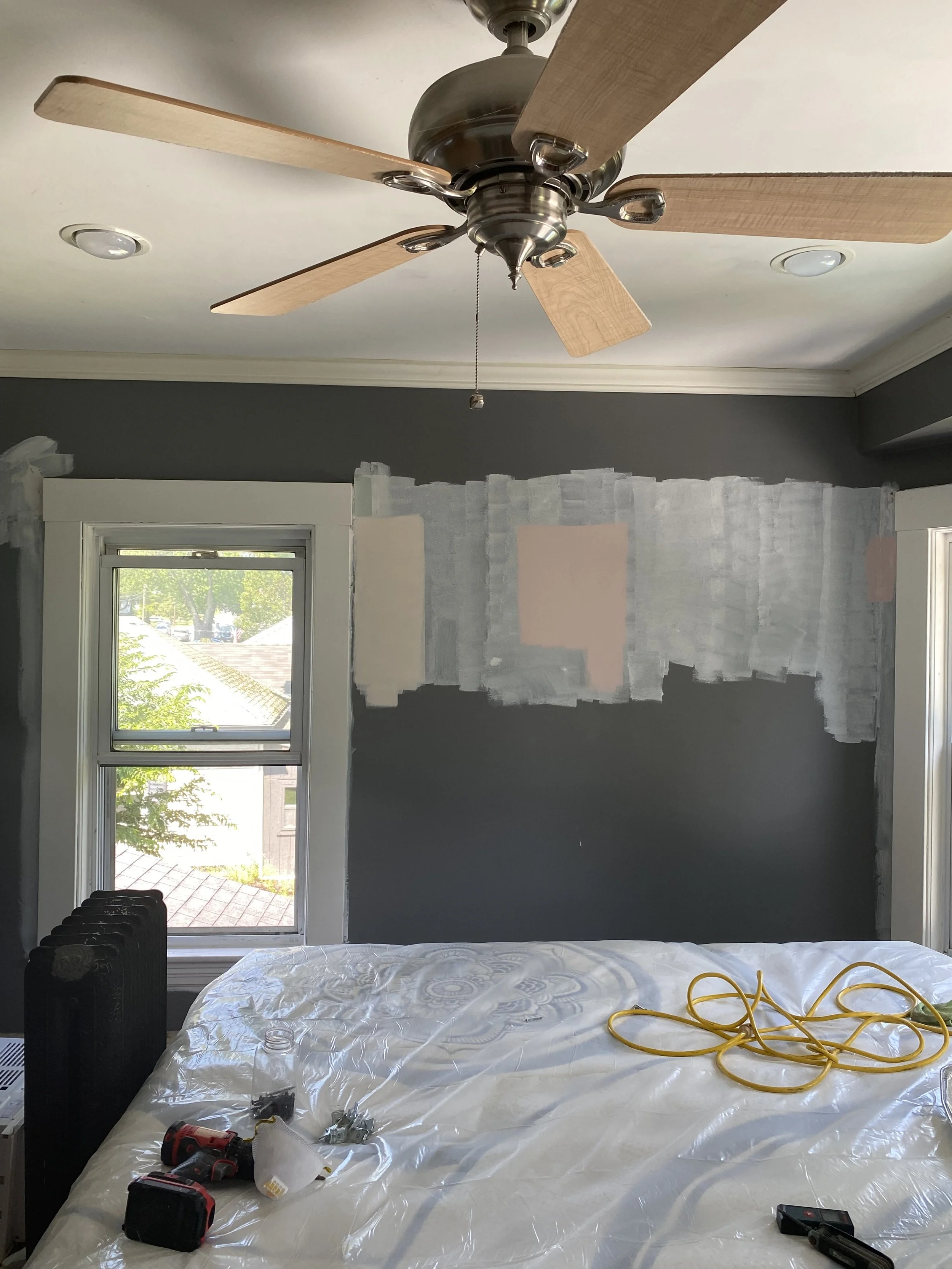 A bedroom undergoing renovation with unpainted patches on the wall, a window showing trees outside, a bed covered with a plastic sheet, and tools including a drill, nails, a yellow extension cord, and small objects on the bed. There's a ceiling fan a