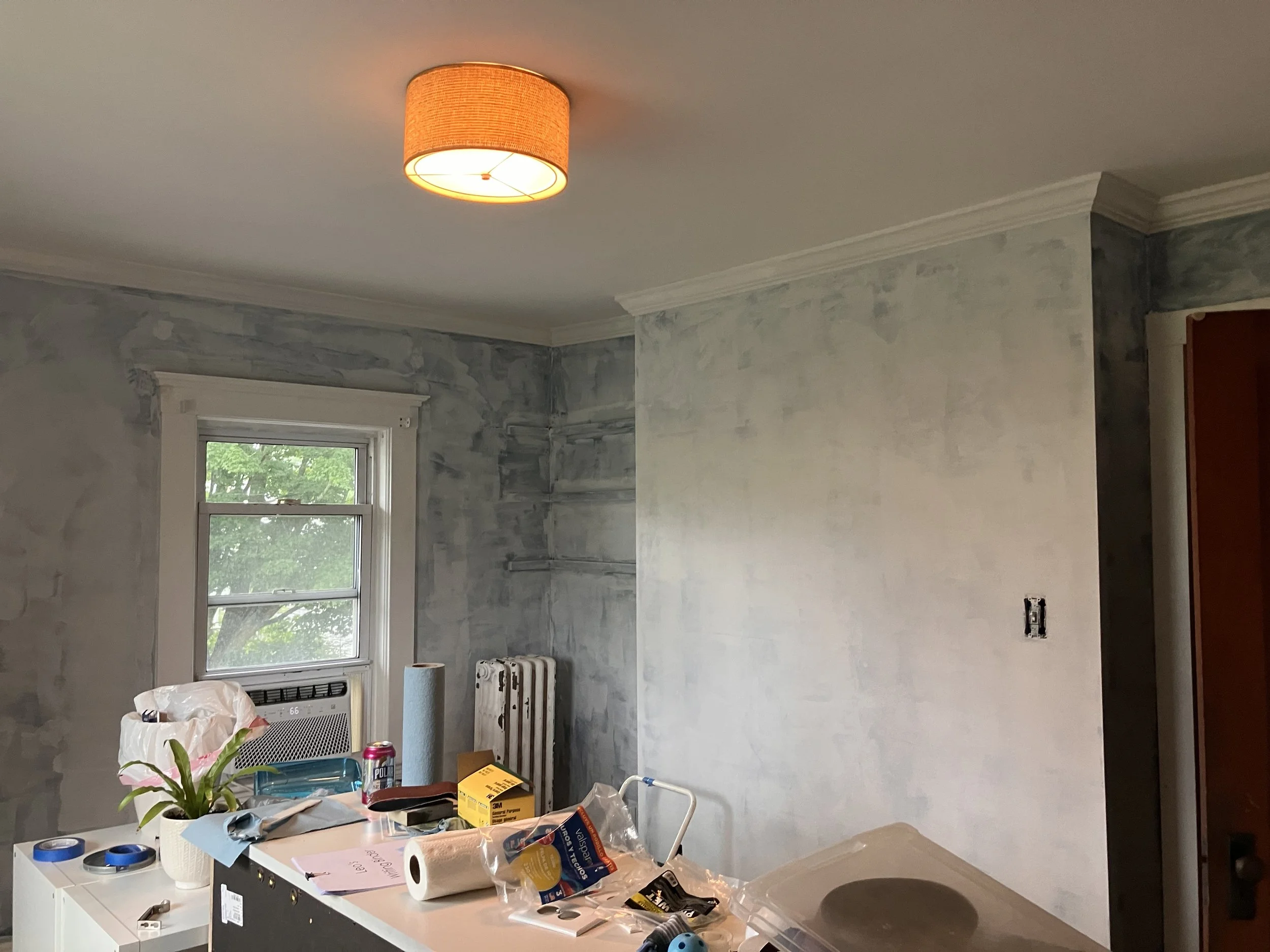 A room under renovation with unpainted walls, a window, a ceiling light with an orange shade, and a table cluttered with various items including a potted plant, a can, a roll of paper towels, and miscellaneous supplies.