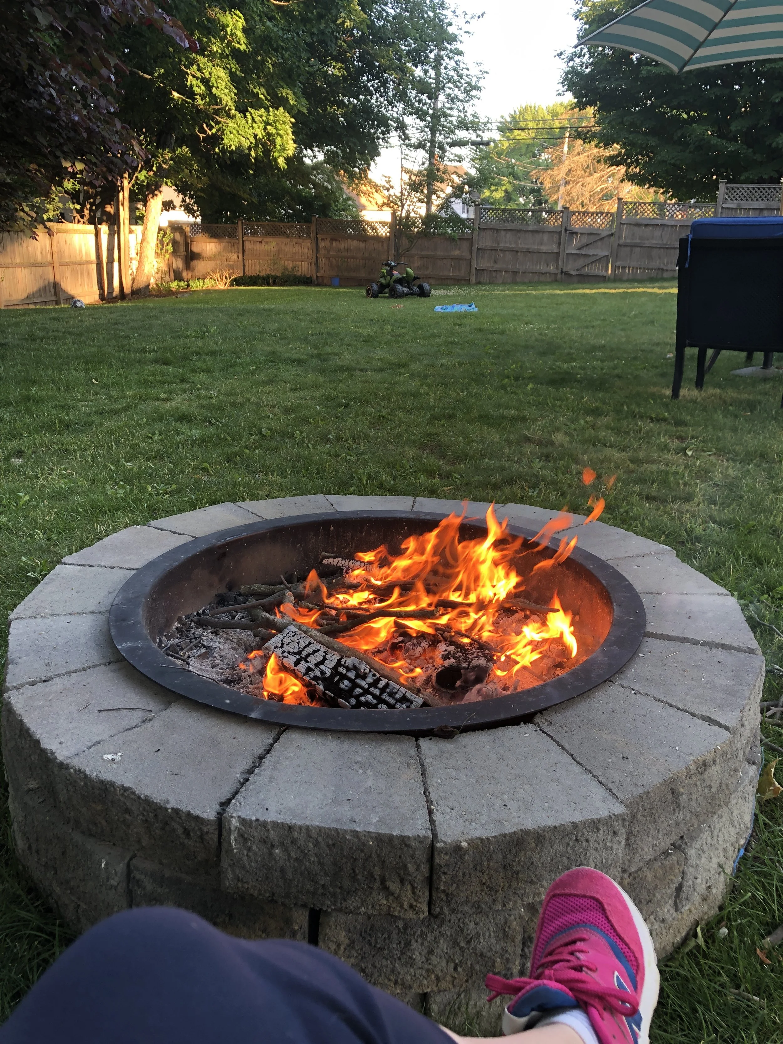 Backyard with a fire pit, grass, trees, a children's toy car, and outdoor furniture, taken in the evening.