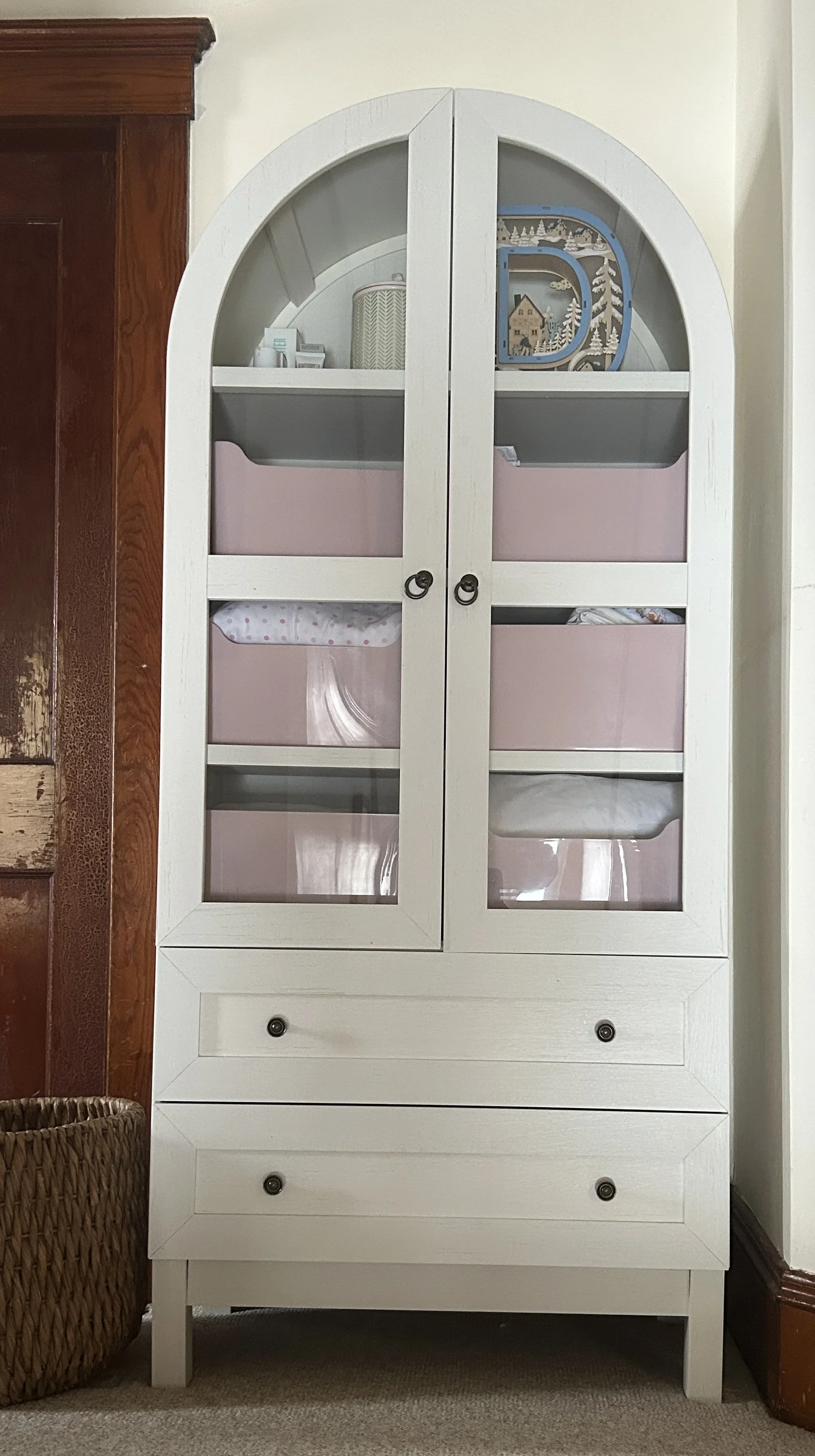 White cabinet with glass doors on top and two drawers at the bottom, placed against a wall next to a wooden door. Inside the cabinet, there are pink storage bins and some decorative items on top.
