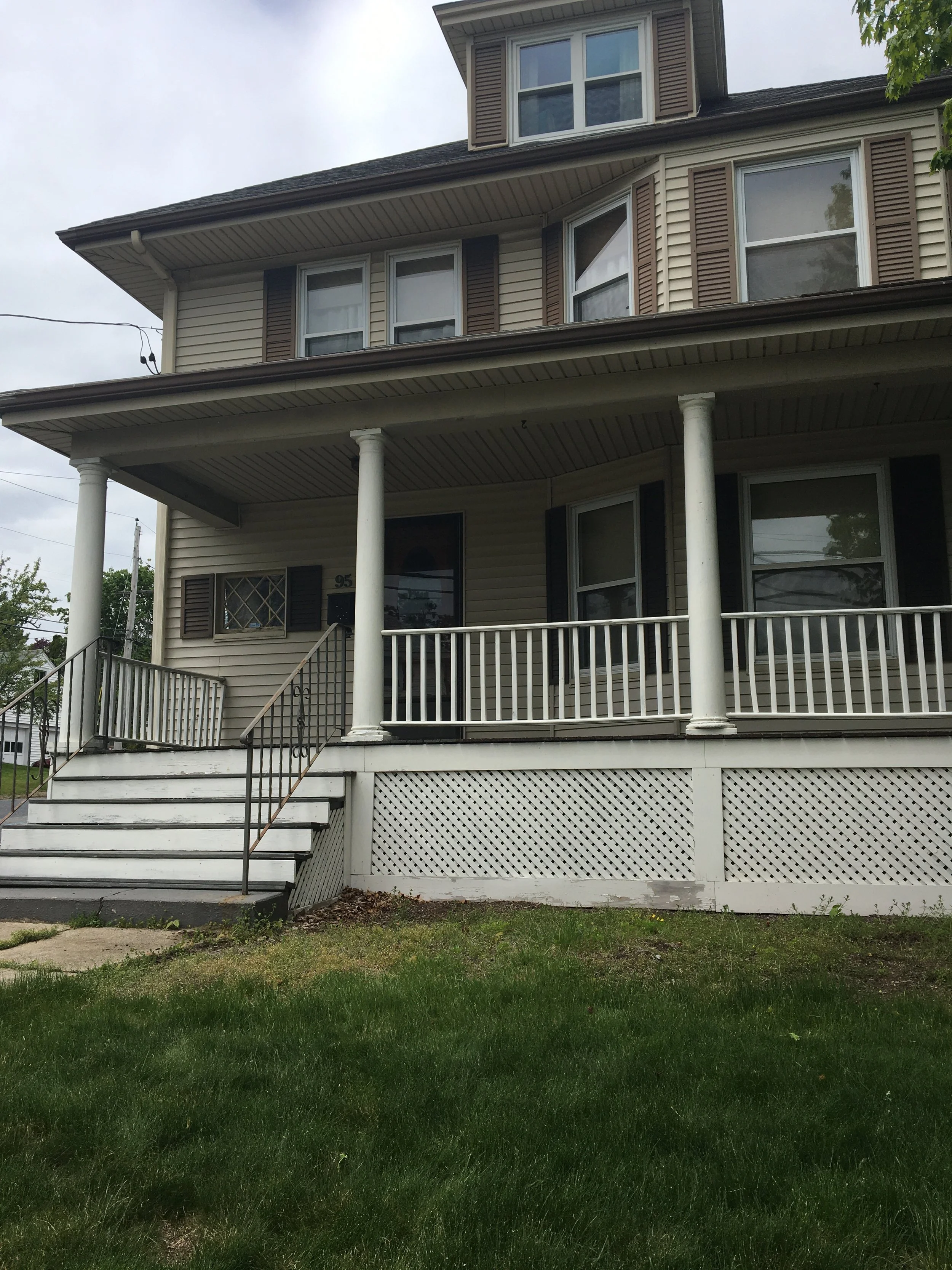 A three-story house with beige siding, white railings, and brown shutters on its windows, featuring a front porch and steps leading up to the entrance.