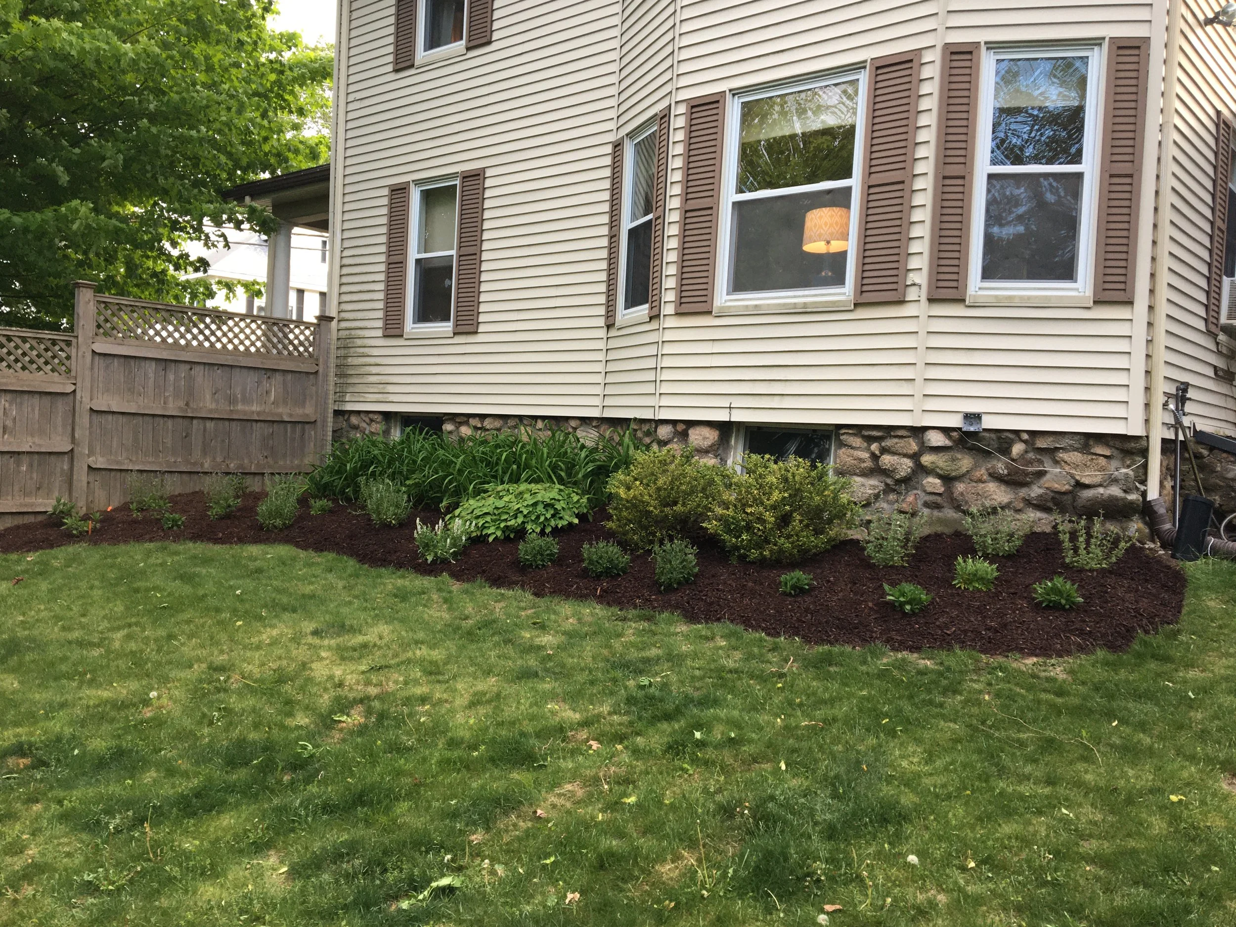 Side view of a house with beige vinyl siding, brown window shutters, and a stone foundation. There is a small garden with bushes and plants along the foundation, and a wooden fence in the background. The lawn is green and well-maintained.