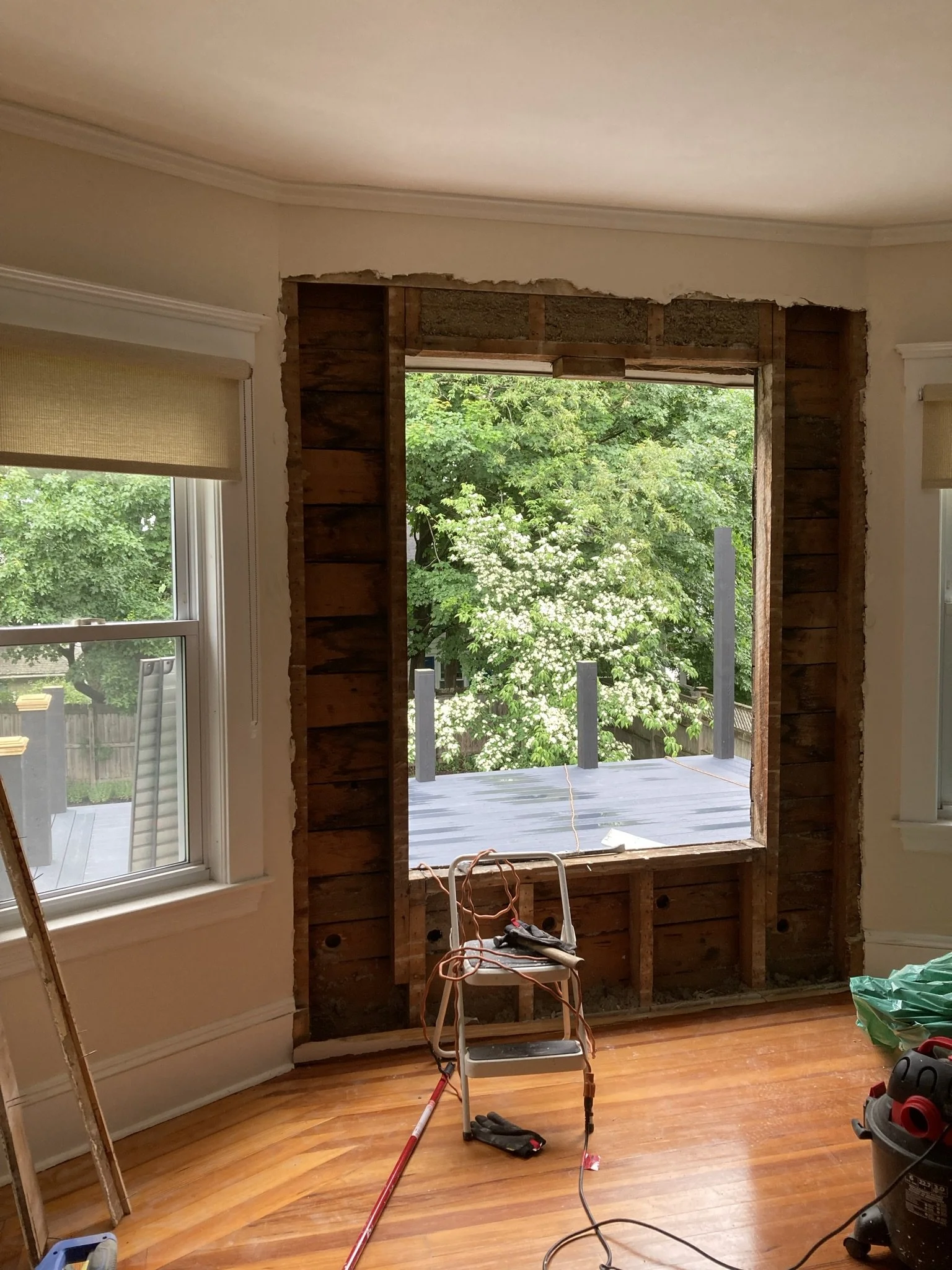 Interior of a room under renovation with a large window opening, exposed wooden framing, and construction tools on the floor.
