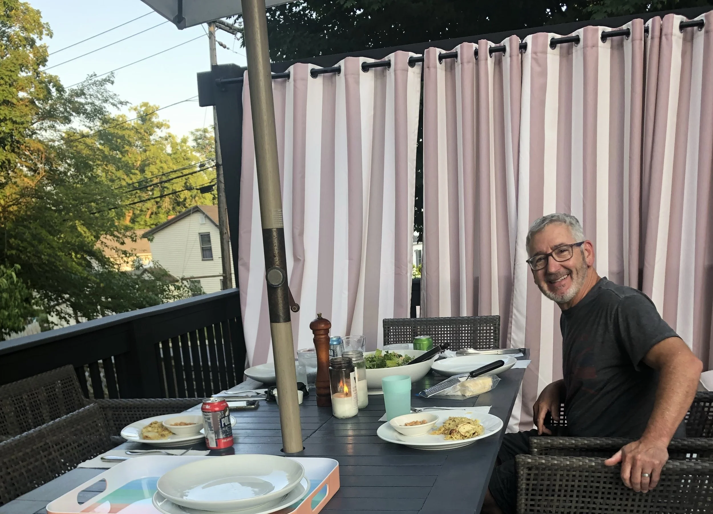 A man with gray hair and glasses smiling and sitting at an outdoor dinner table with plates of food, drinks, and salt and pepper shakers, on a balcony with pink and white striped curtains, surrounded by trees and houses in the background.