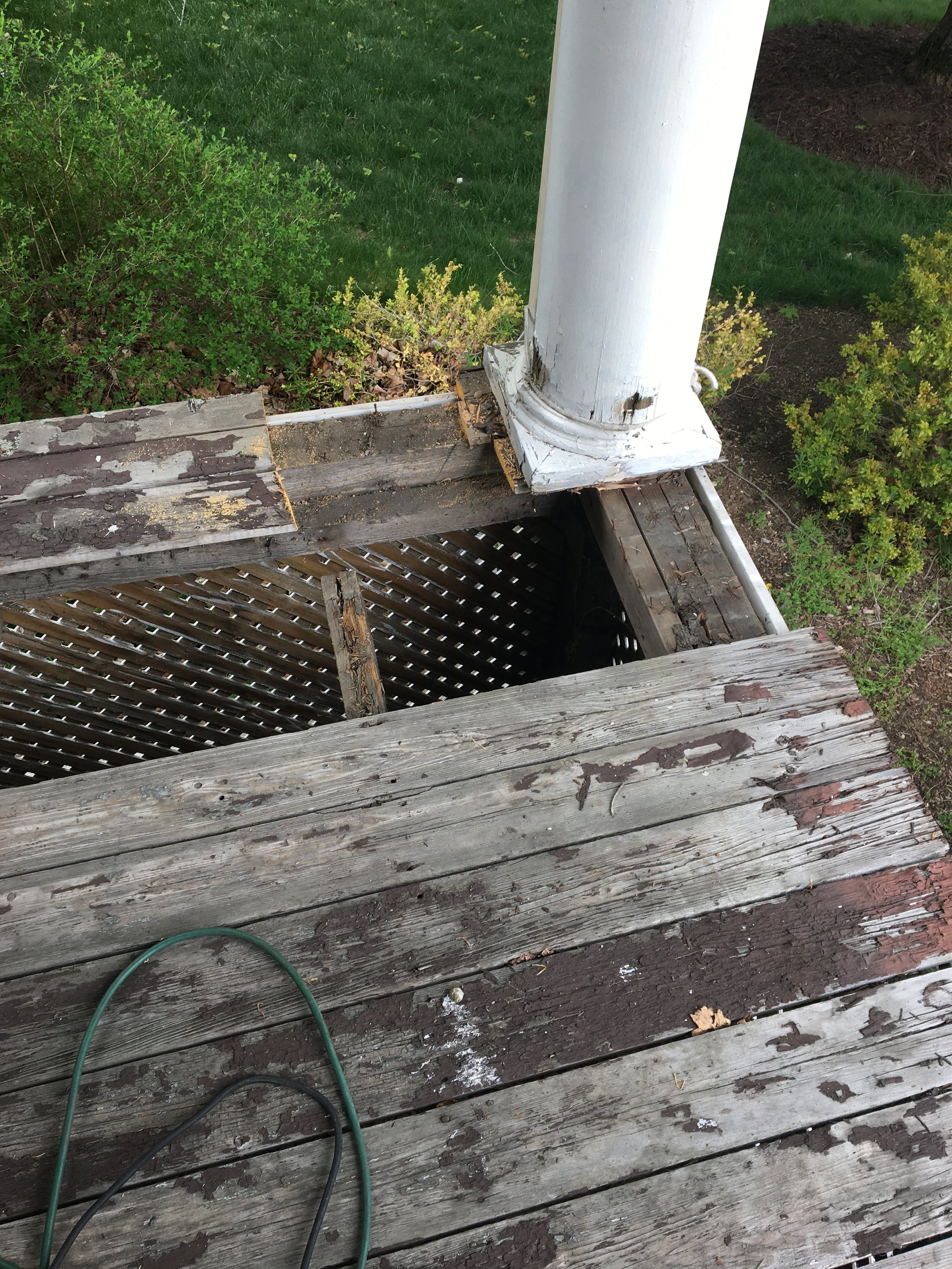 A weathered wooden deck with a hole where a square section has been removed, revealing the lattice underneath. The deck has peeling paint and some leaves on it. There is a white post attached to the corner of the deck, and some green and black electr