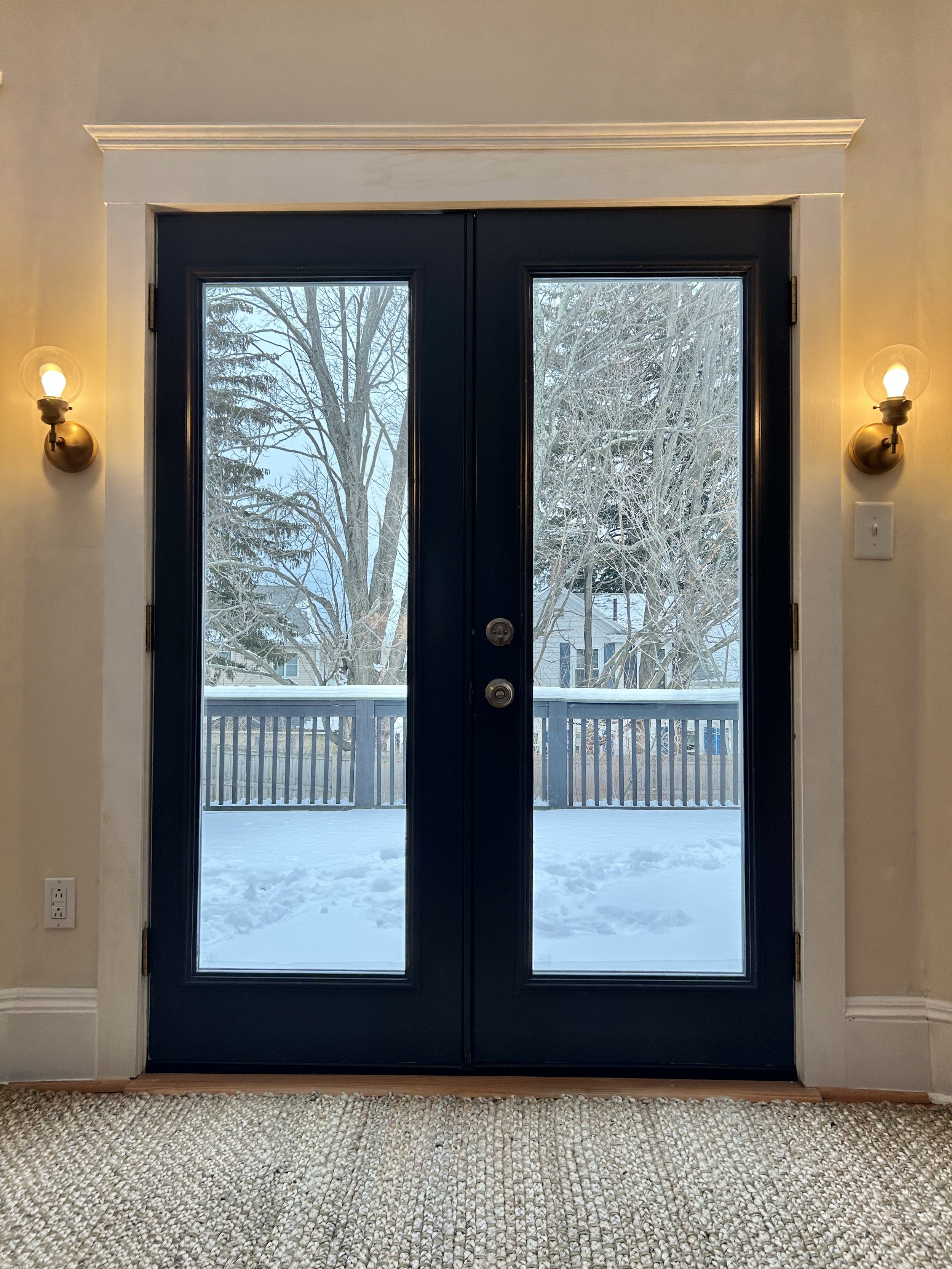 Interior view of a door with glass panels showing a snowy yard with trees and houses outside, flanked by beige walls and wall-mounted light fixtures on each side.