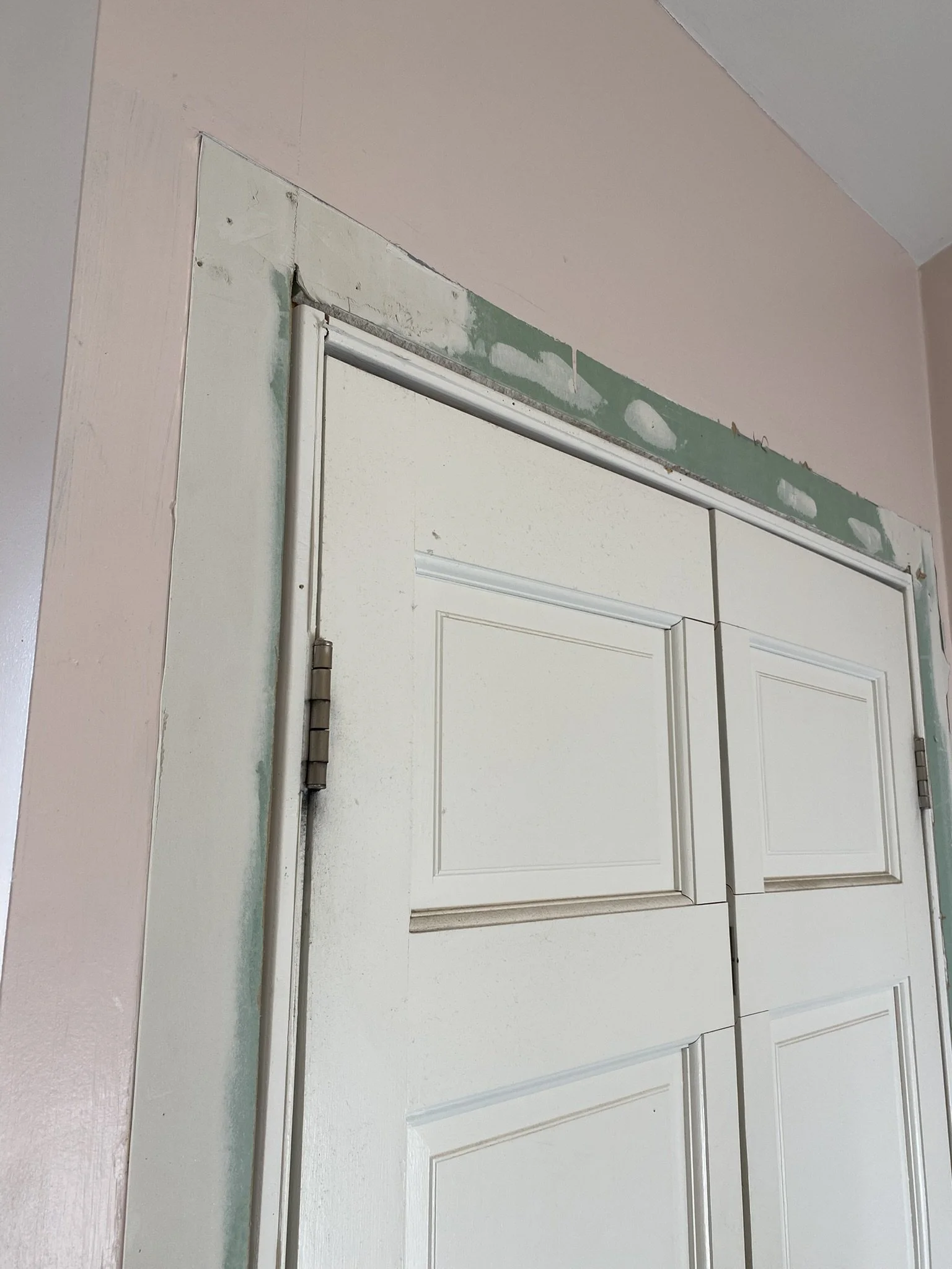 Close-up of a white double door with missing trim at the top, showing unfinished green drywall and some wall patching in progress.