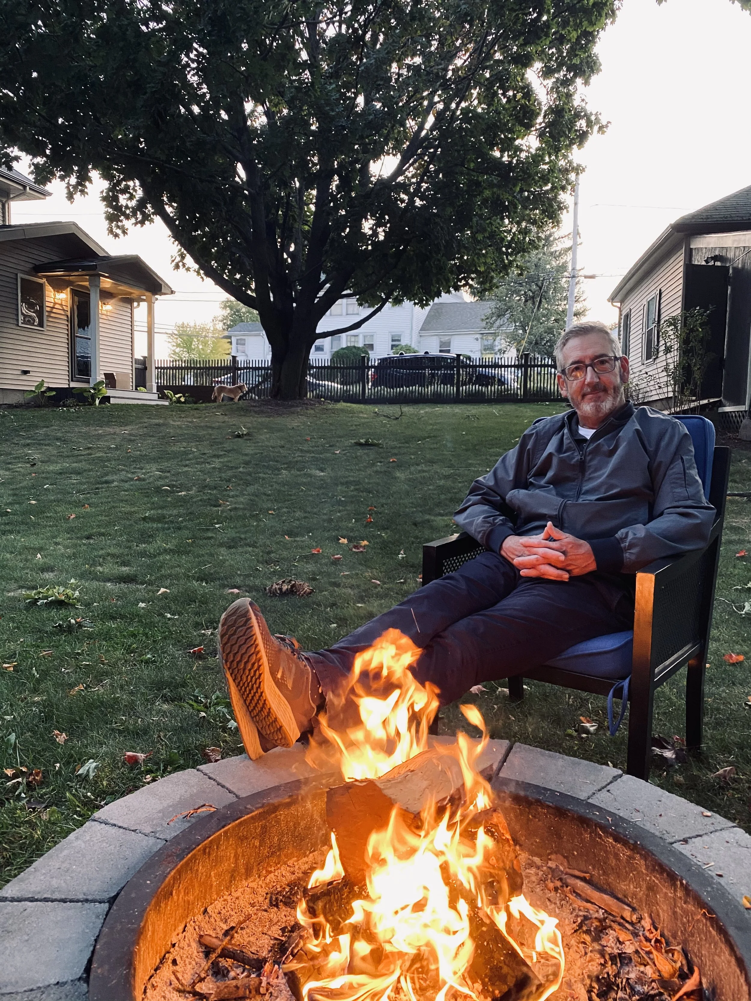 A man with glasses and a beard sitting with his feet in a fire pit, smiling, in a backyard during dusk. There is a tree and houses in the background.