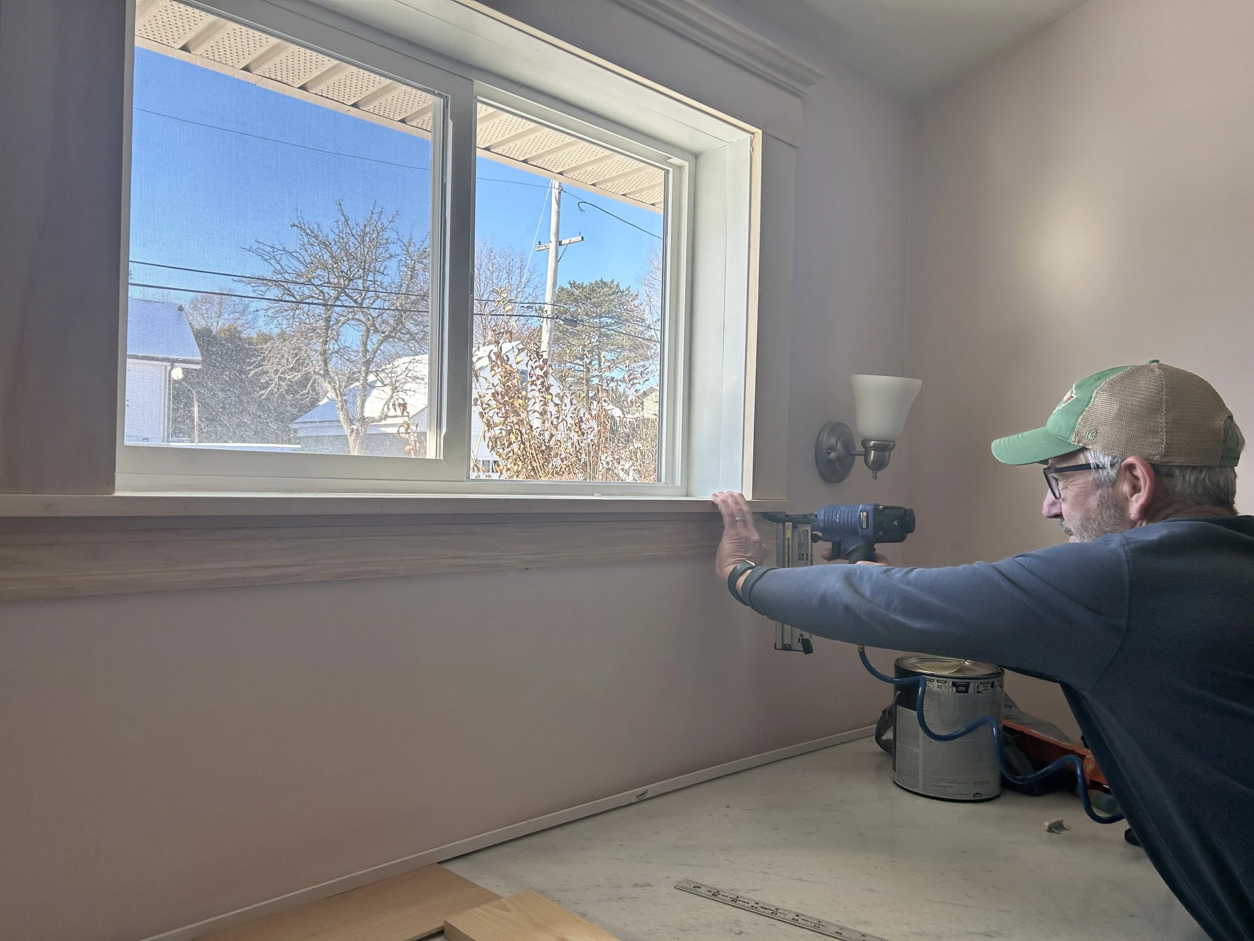 A man wearing a cap and glasses is installing or repairing a window in a room, using a cordless drill on the window frame. Outside the window, there is a clear view of trees and rooftops under a blue sky.