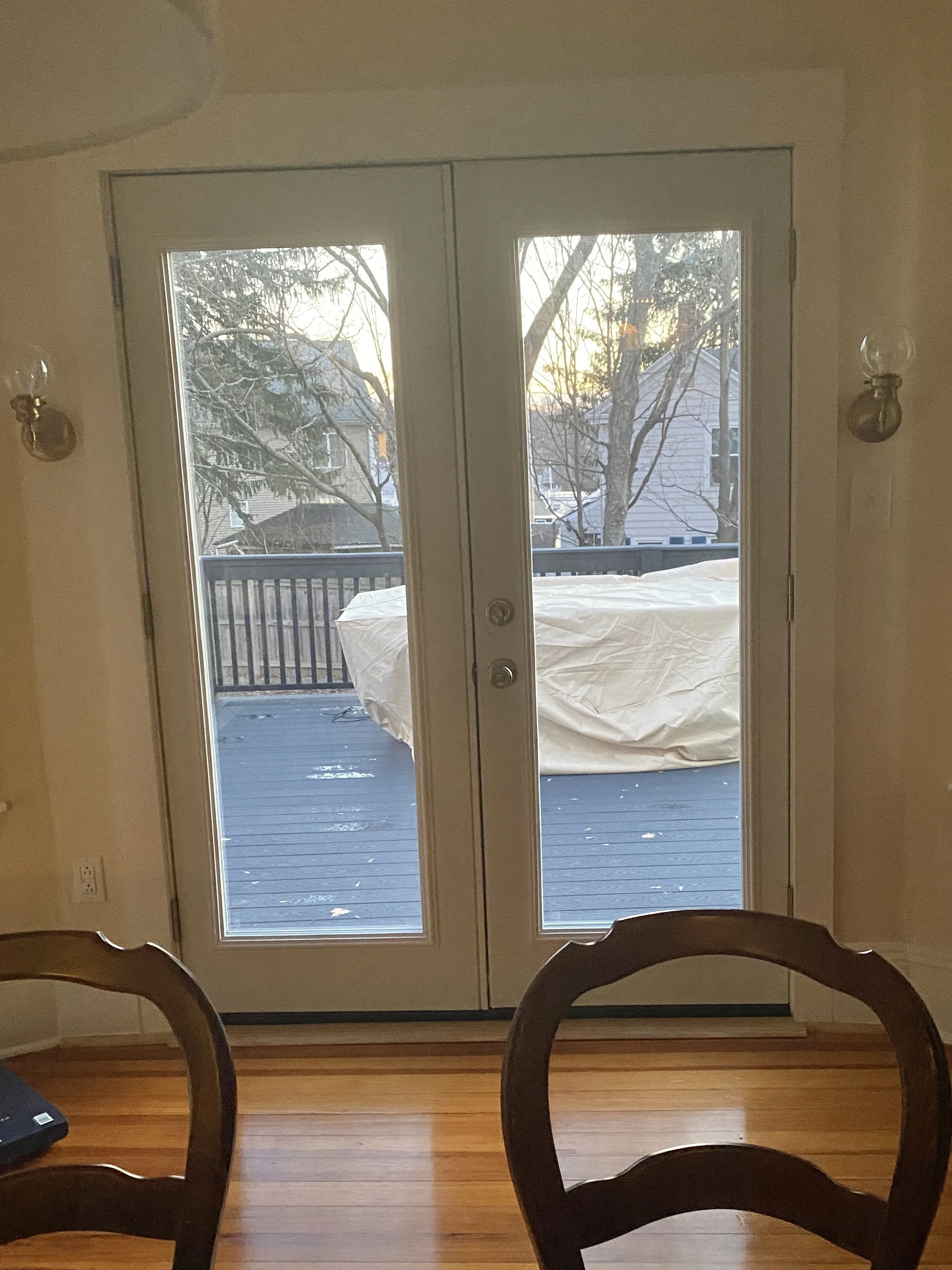 View of a deck through glass double doors, with a covered outdoor furniture piece and leafless trees in the background, taken during winter.