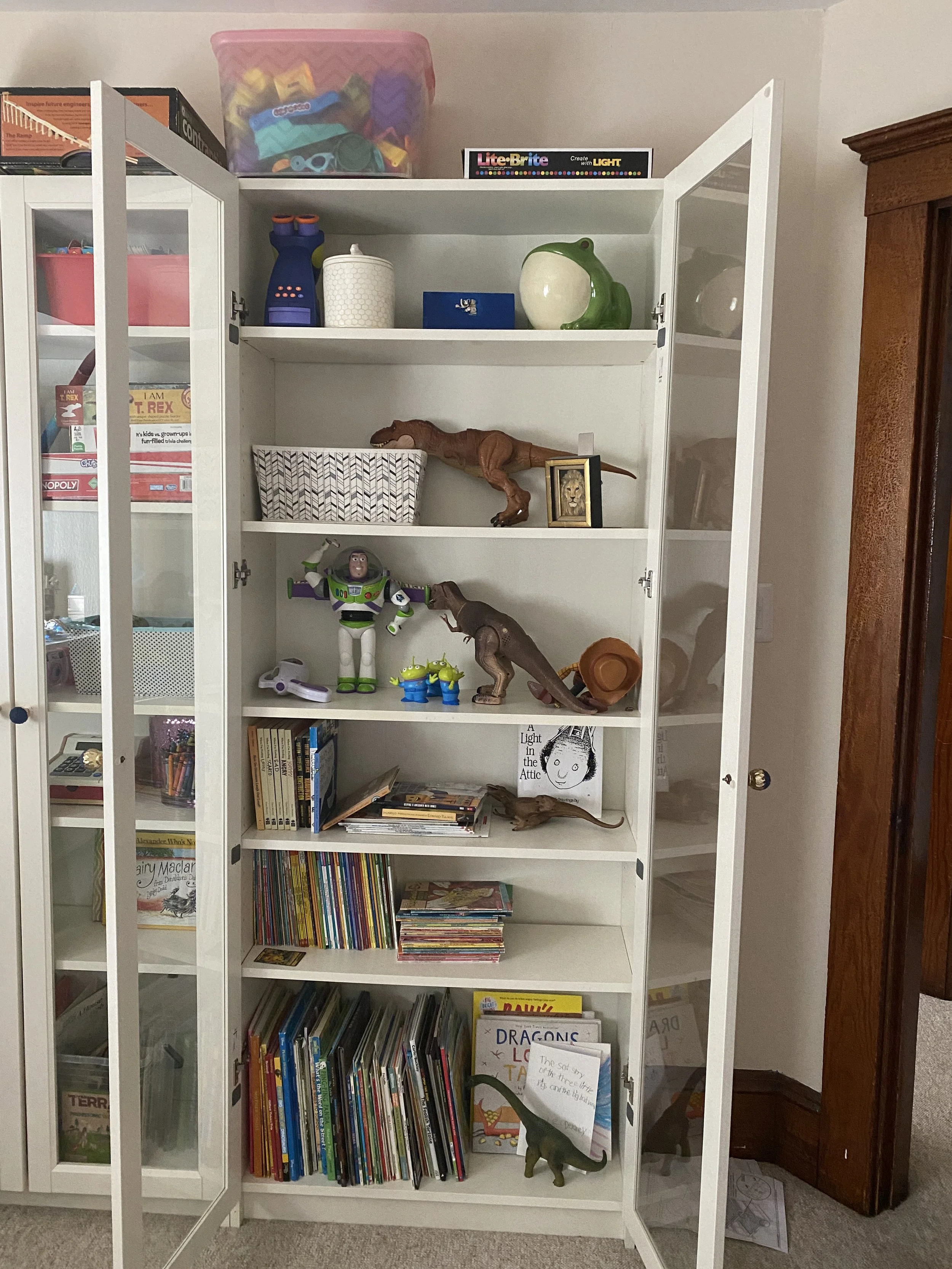 A white bookshelf with glass doors partially open, filled with toys, books, and decorative items. The top shelf has containers and a box of Lite-Brite. Middle shelves hold dinosaur toys, Buzz Lightyear, and various children's books. The right side ha