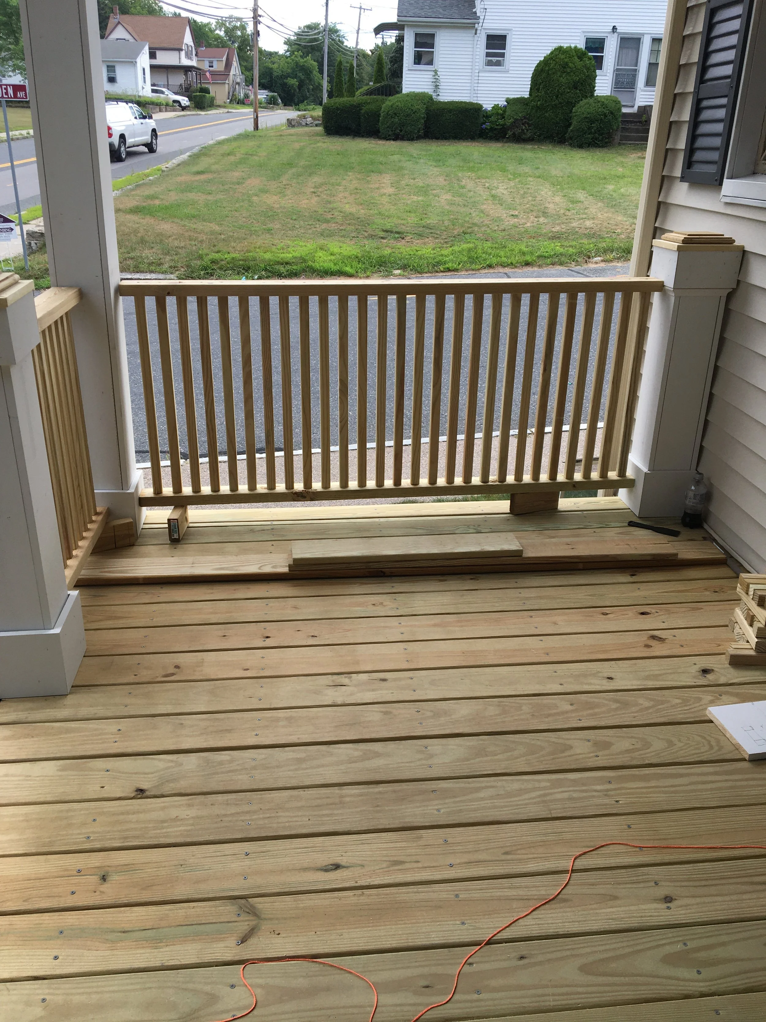 View of a newly constructed wooden deck with a railing, facing a grassy area and residential houses in the background, during daytime.