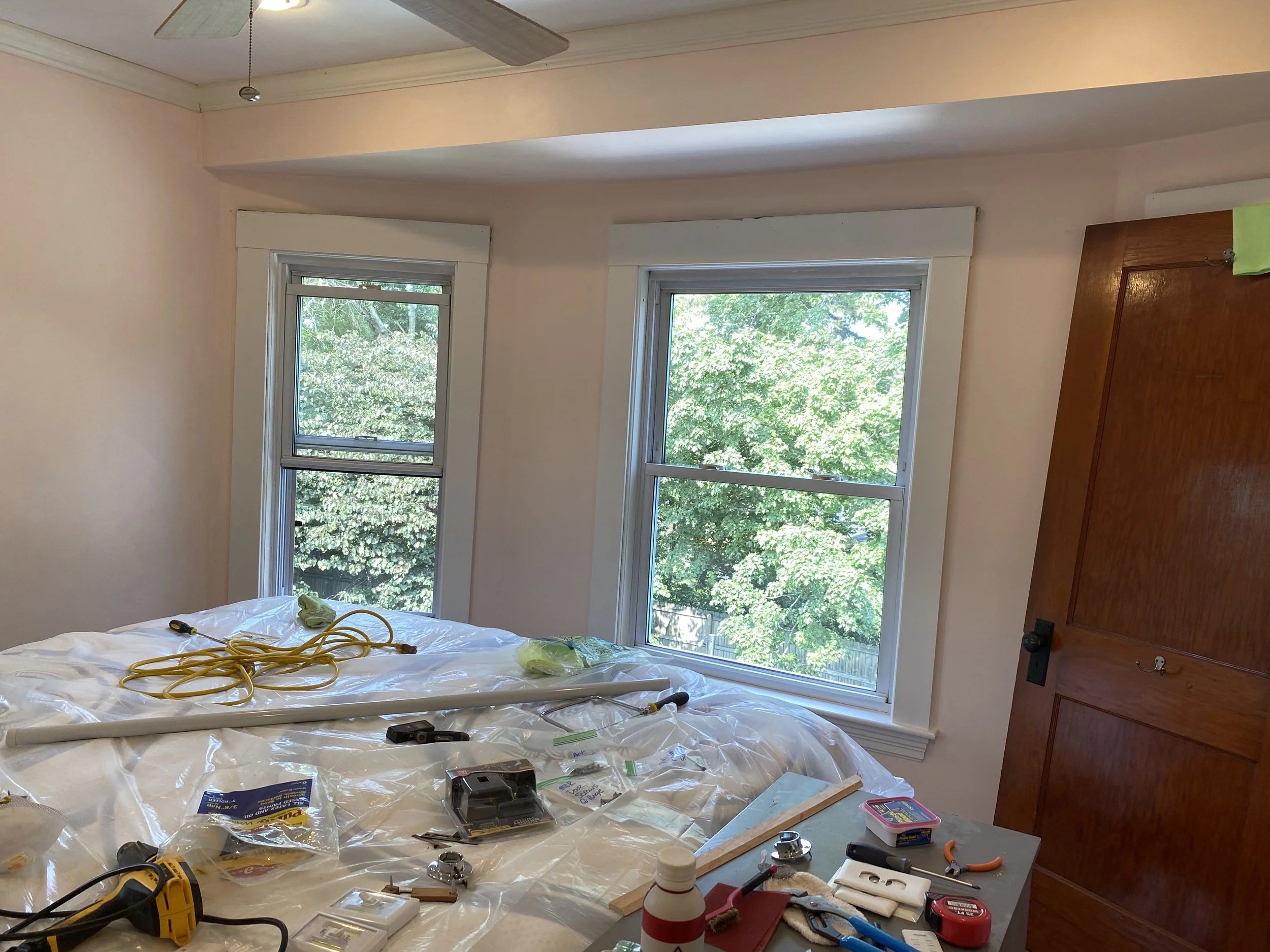 A room with two windows showing green trees outside. The room contains construction tools and supplies, including a yellow cord, a black tool, a white pipe, a level, and various small items on a covered surface, indicating ongoing home renovation or 