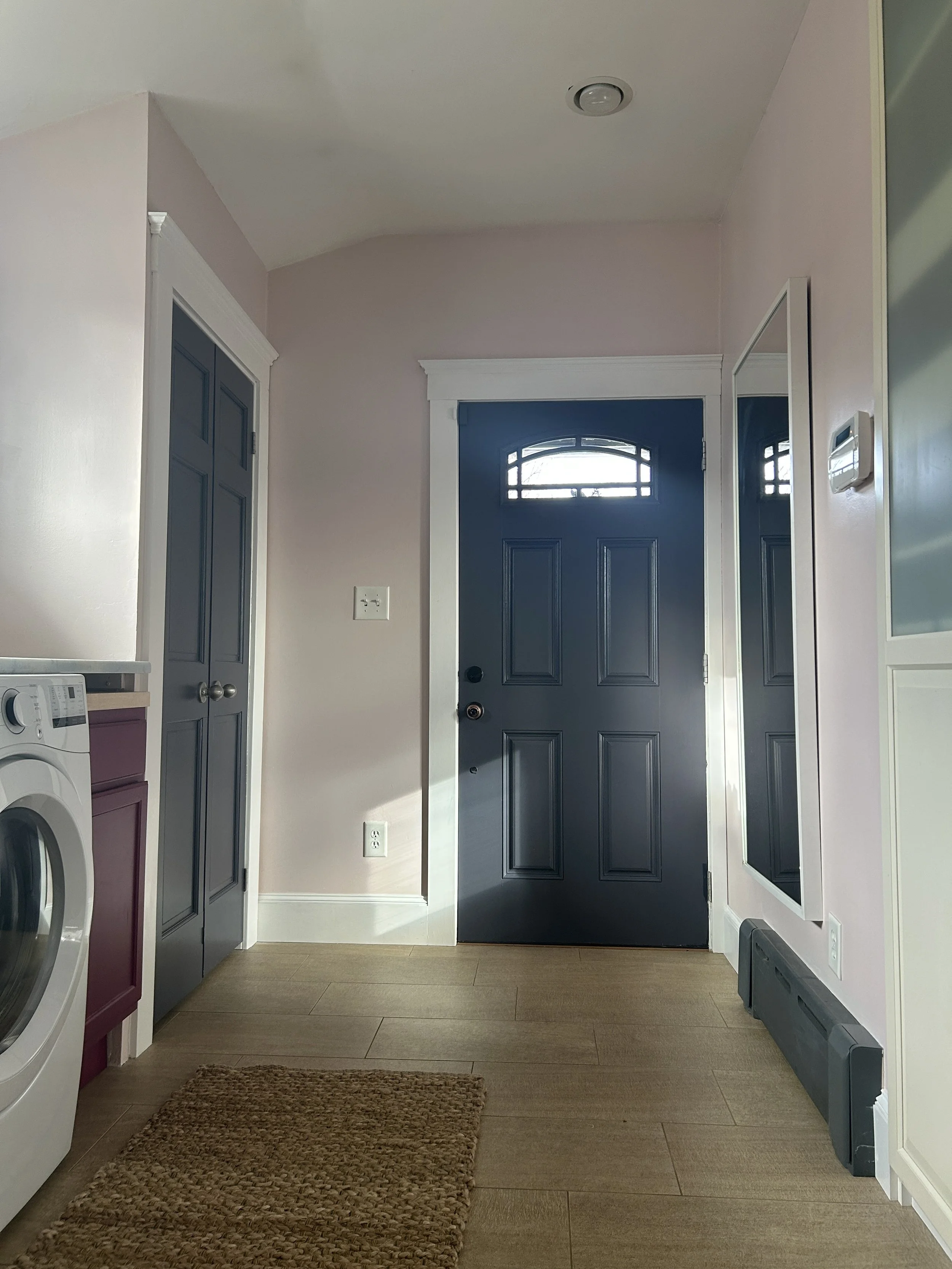 Interior view of a laundry room with a dark blue front door, a mirror on the right wall, a washing machine on the left, a small cabinet, and a brown woven rug on the wooden floor.
