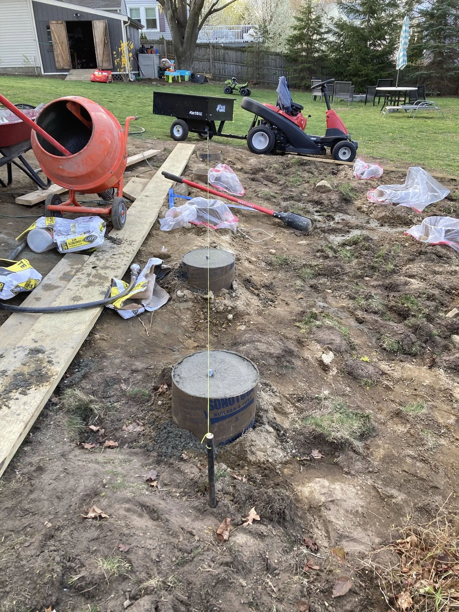 A backyard under construction with cement pillars, construction tools, and equipment scattered around. There is a concrete mixer, a wheelbarrow, and a small toy tractor. The area appears prepared for a new structure or project, with some plastic cove