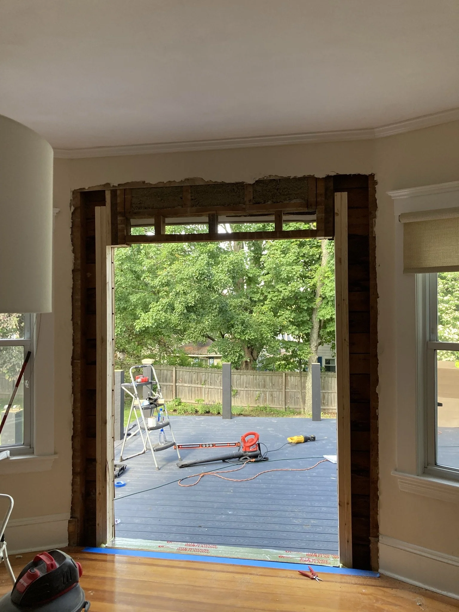 Room undergoing renovation with a large open doorway to an outdoor deck, construction tools and equipment including a ladder, leaf blower, drill, and extension cord, with trees and a wooden fence outside.