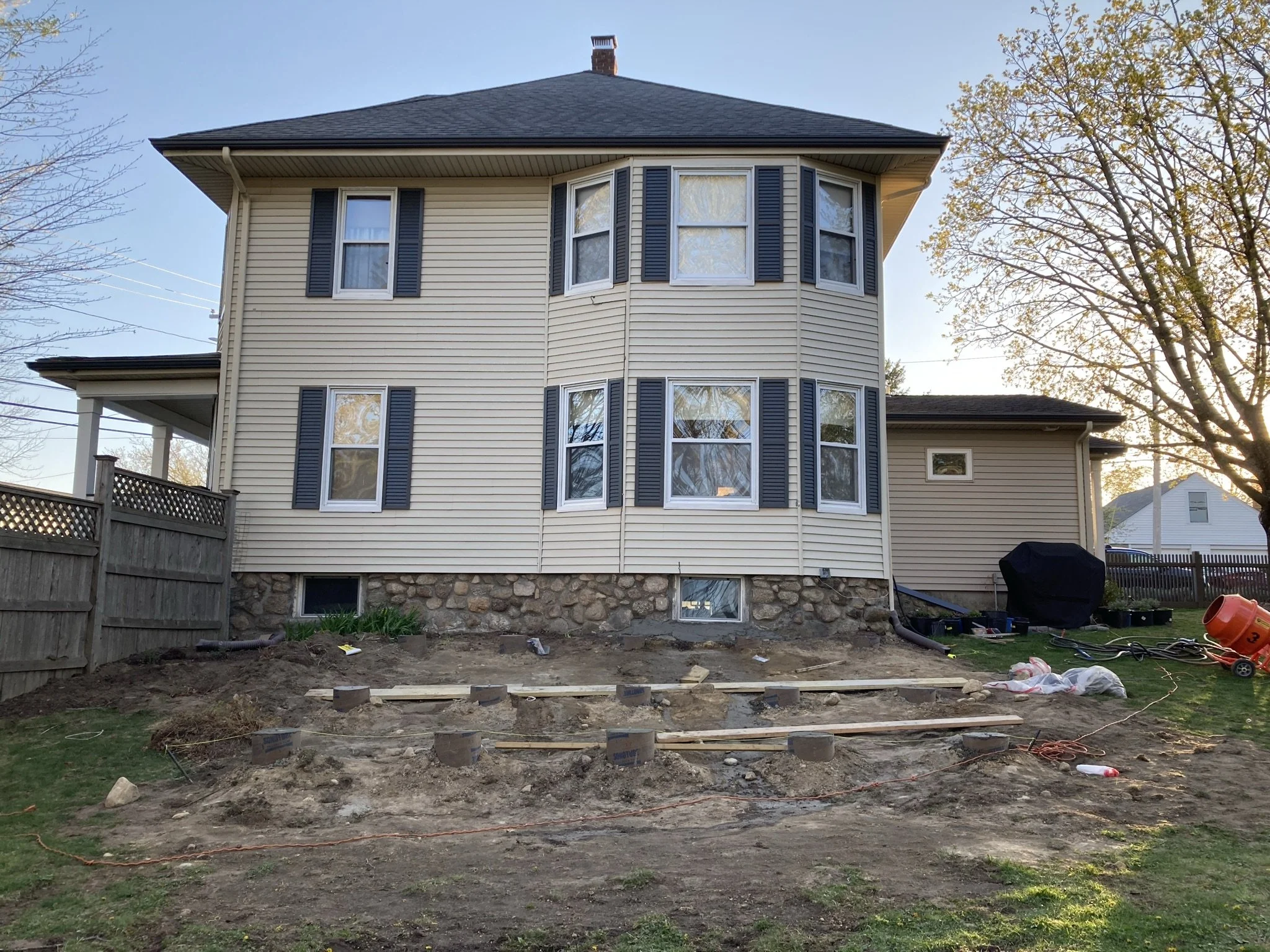Backyard of a house under construction, with dirt and wooden planks in the foreground, windows with dark shutters, and trees on either side under a clear sky.
