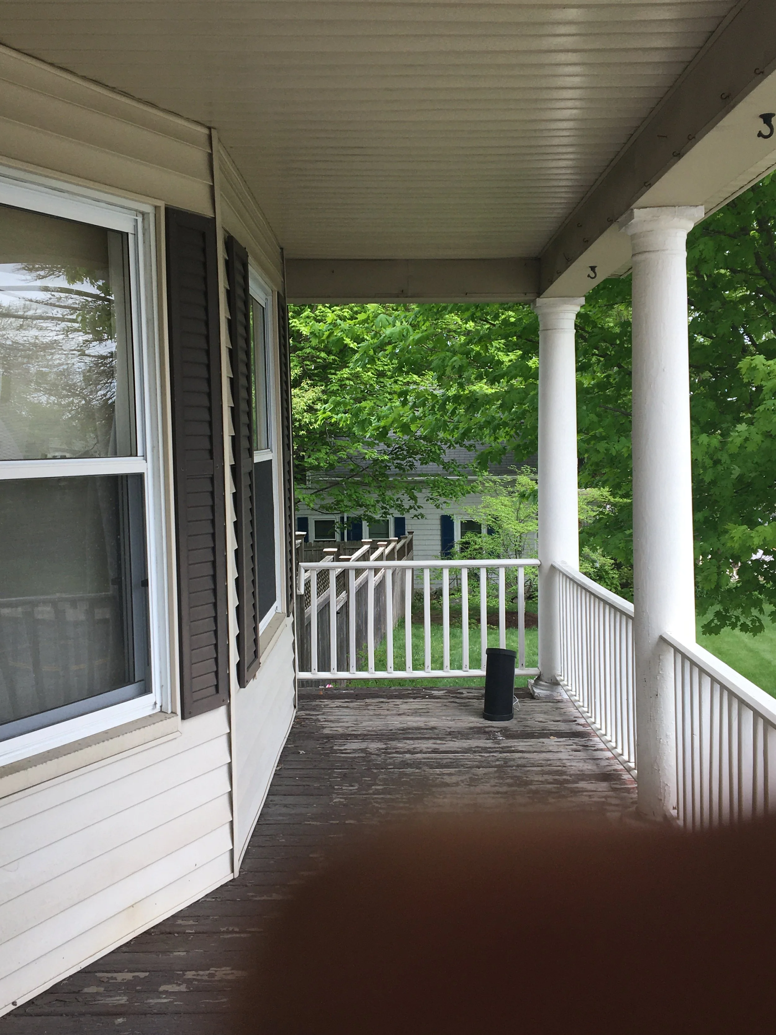 View of a porch with a weathered wooden floor, white railings, two white columns, and part of a house with beige siding and black shutters. Green trees and neighboring houses are visible in the background.