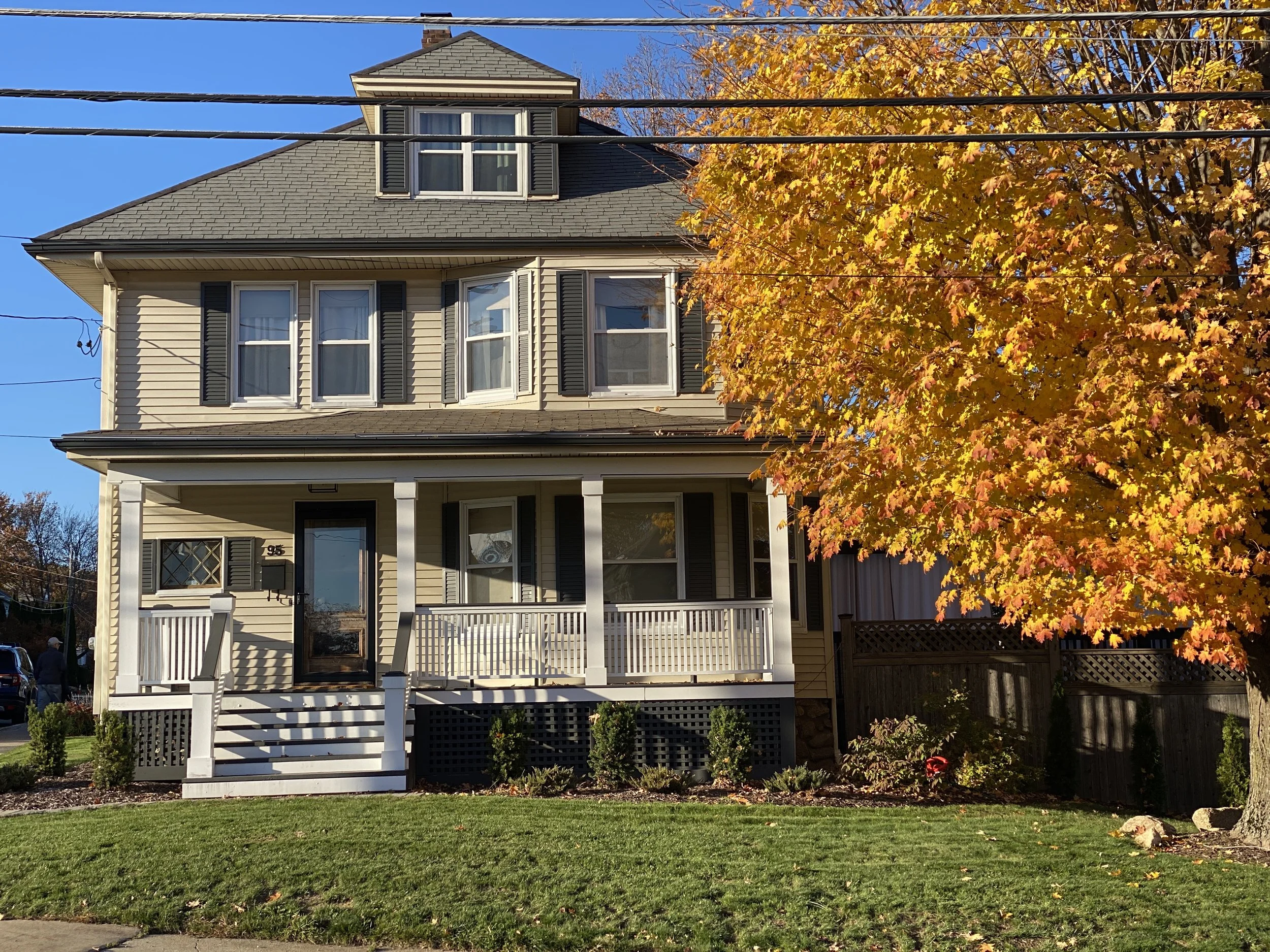 A three-story beige house with black shutters, a front porch, and a staircase, situated on a grassy lawn with a large orange autumn tree and a blue sky in the background.