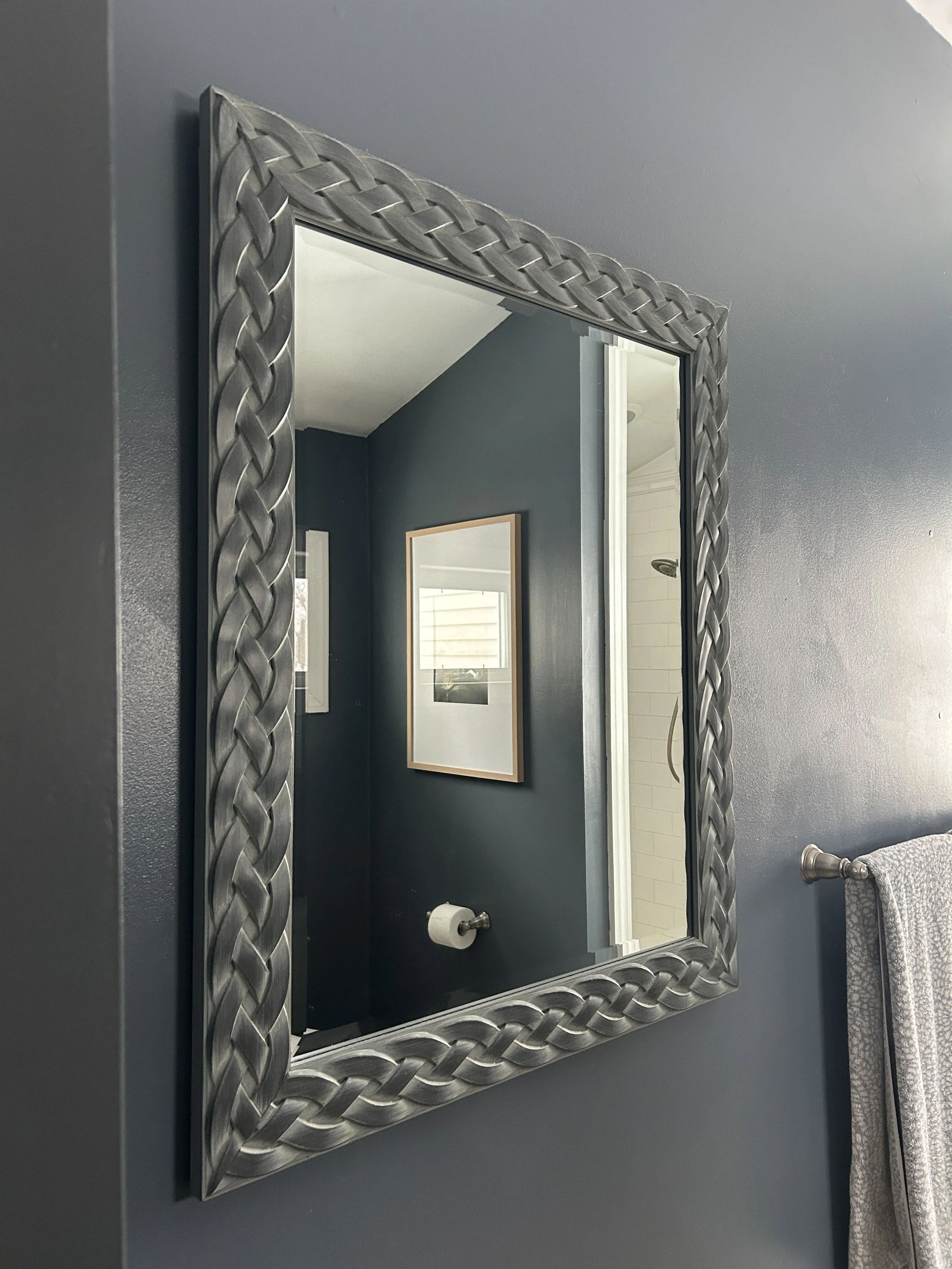 A bathroom mirror with a decorative metal frame mounted on a blue wall. In the reflection, a framed picture, a window, and a shower area are visible.