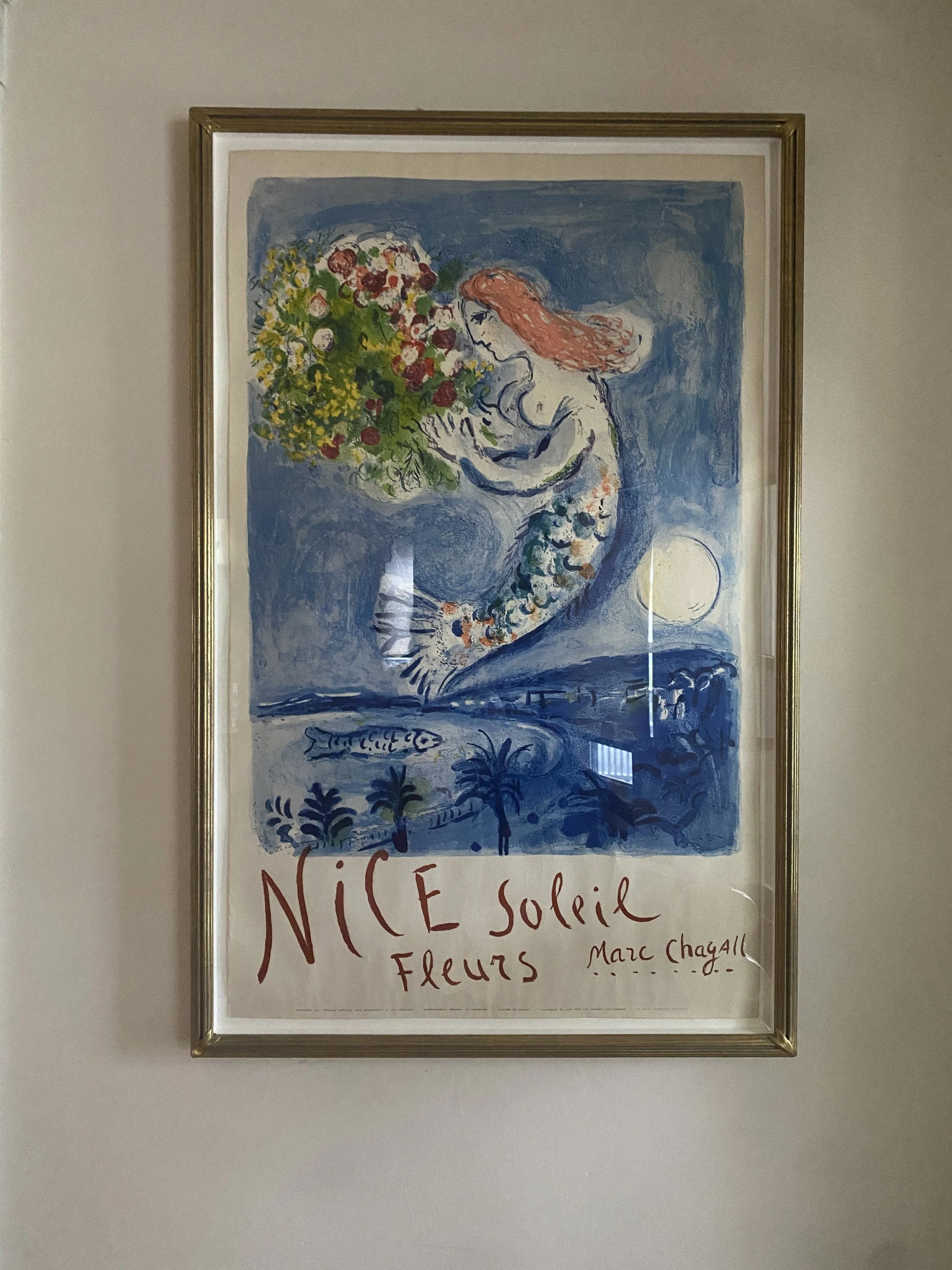 A framed poster of a painting by Marc Chagall featuring a mermaid holding a large bouquet of flowers with a moonlit landscape in the background and the words 'NICE soleil fleurs' written at the bottom.